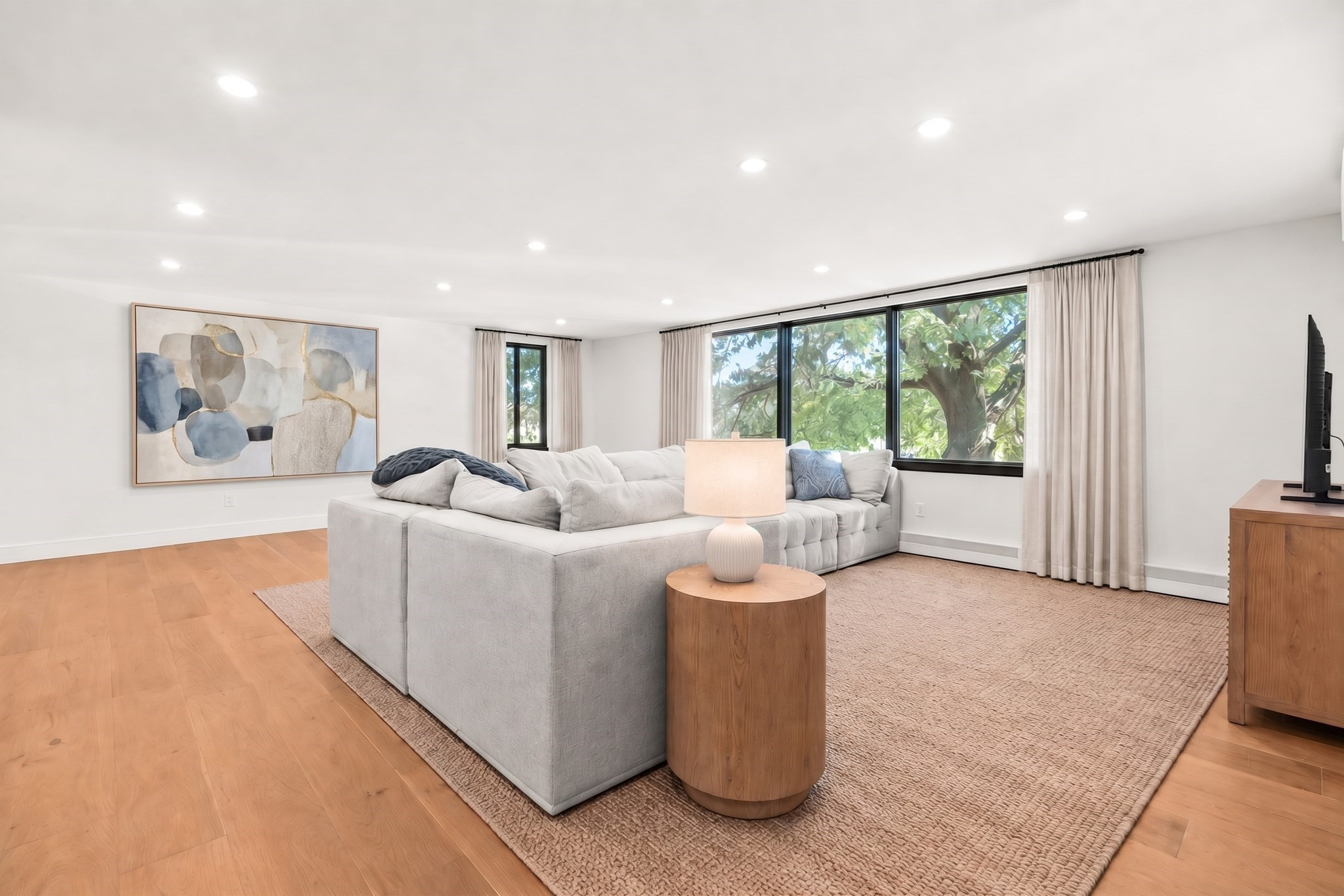 Living room with a light-gray sectional sofa, warm wood floors, and wide windows overlooking leafy trees.