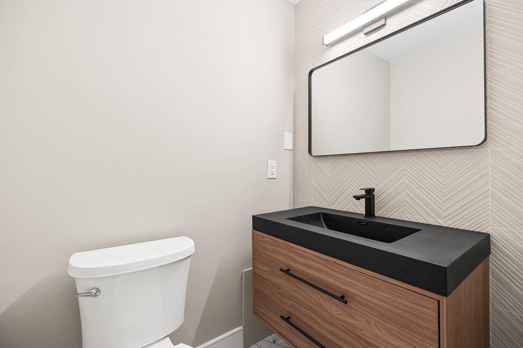 Bathroom with a wood vanity, black countertop, and herringbone tile behind a large mirror.
