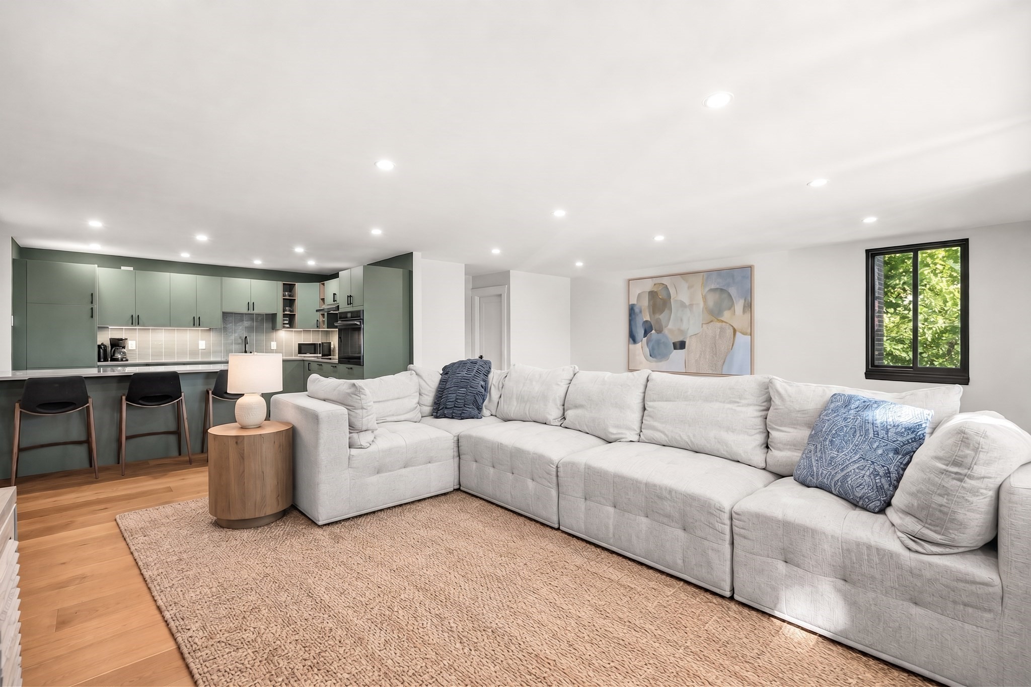 Open-plan living room with a large gray sectional facing a sage-green kitchen.