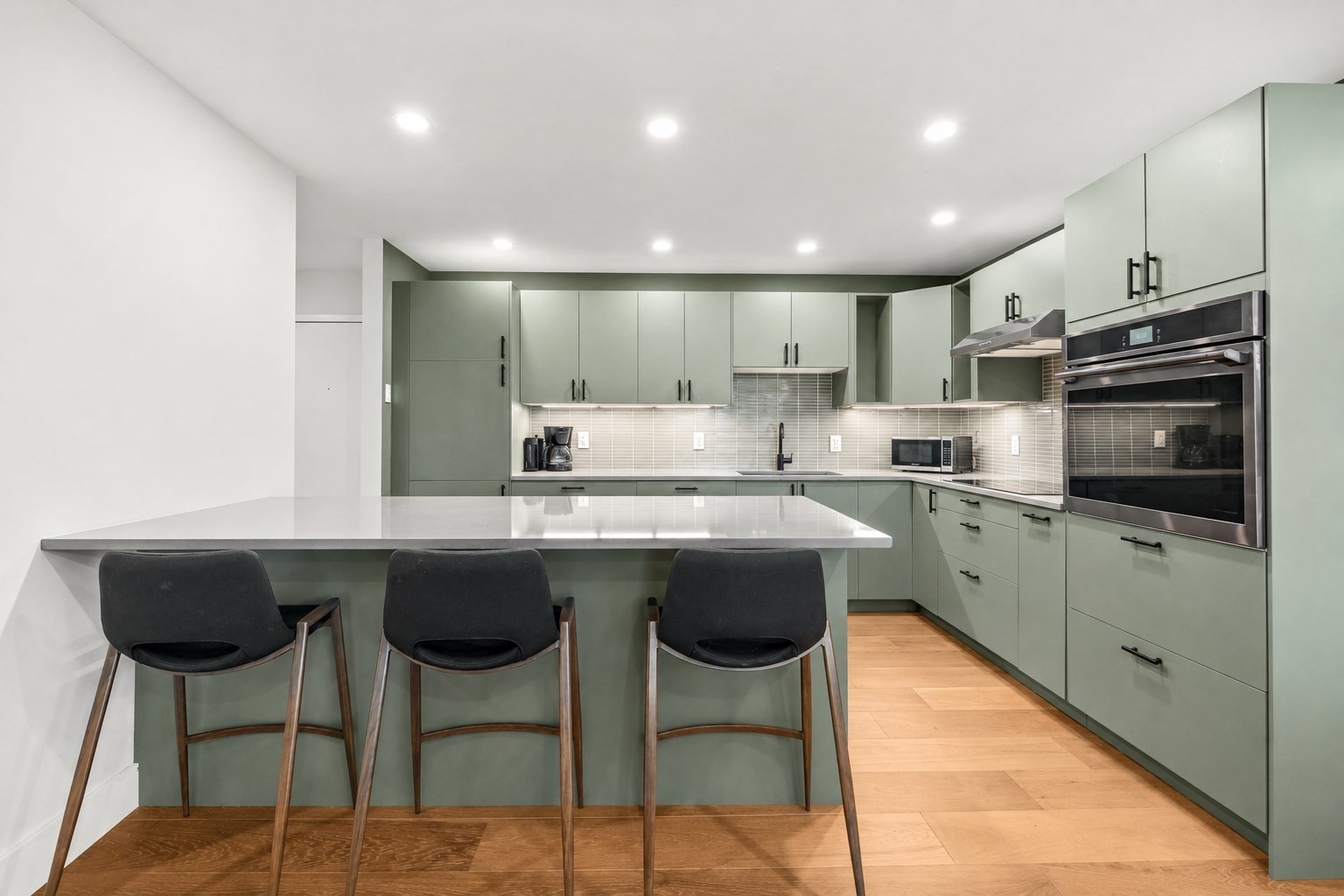 Kitchen with sage-green cabinets, white island, three bar stools, and stainless-steel double oven.