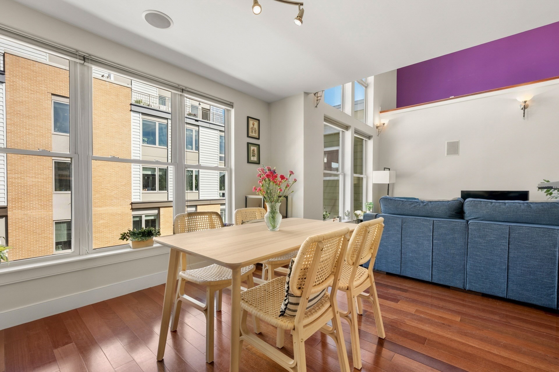 Dining area with wicker chairs and a wood table beside a blue sofa and tall windows.