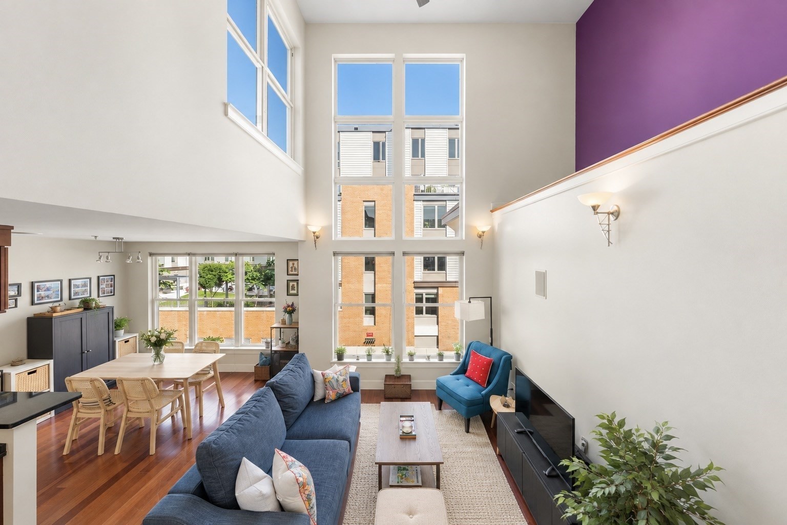 Double-height living room with tall windows, blue seating, hardwood floors, and an adjoining dining area.