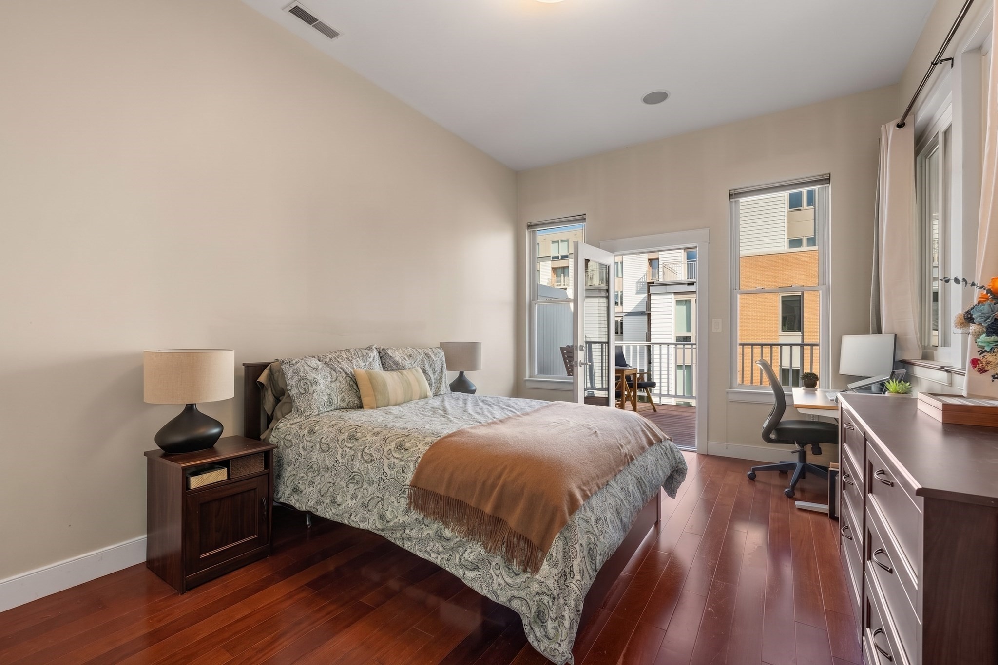 Bedroom with a patterned bed, wood floors, a desk, and an open balcony door.