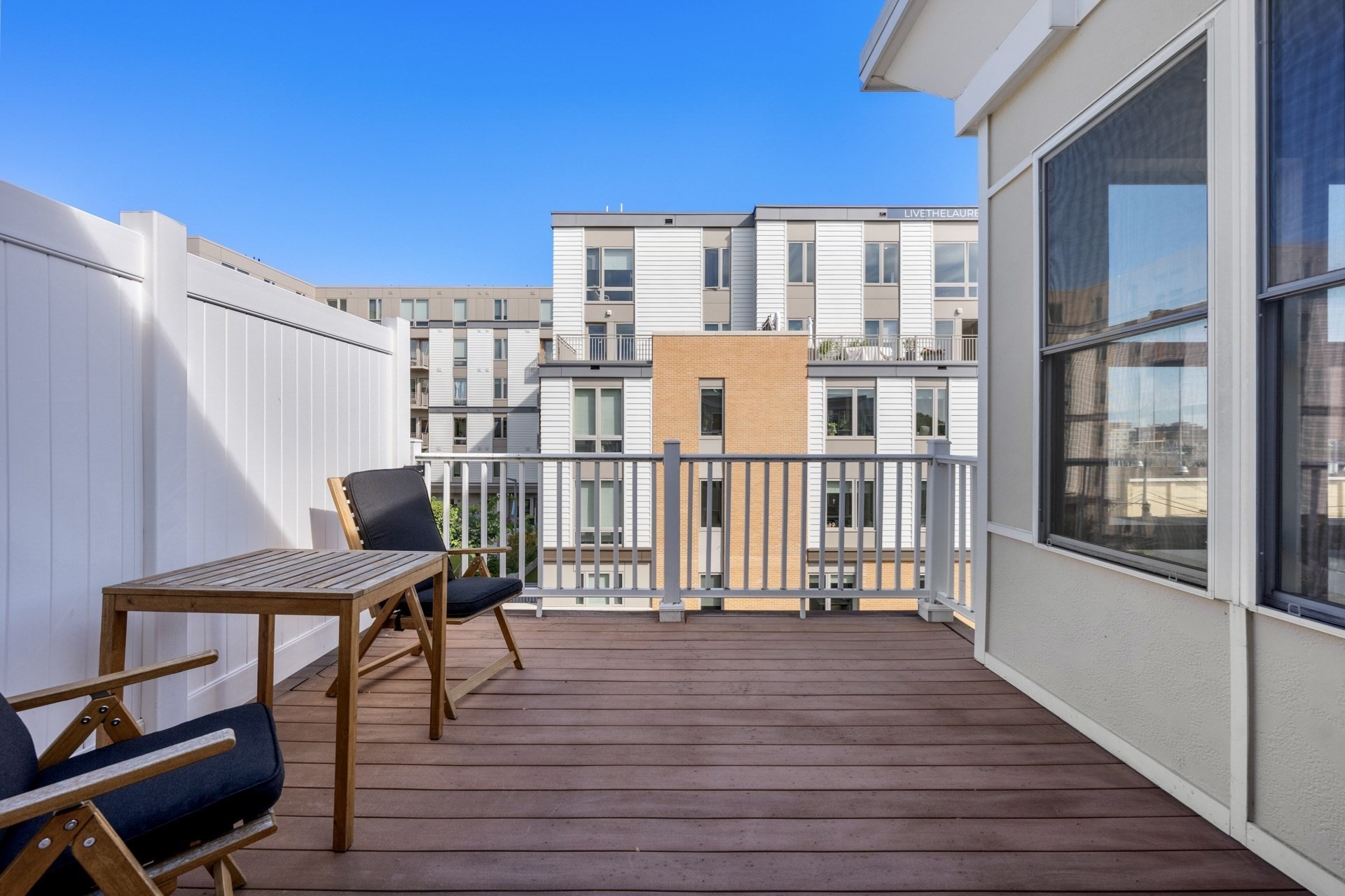 Apartment balcony with wooden table and cushioned chairs overlooking a modern residential building.