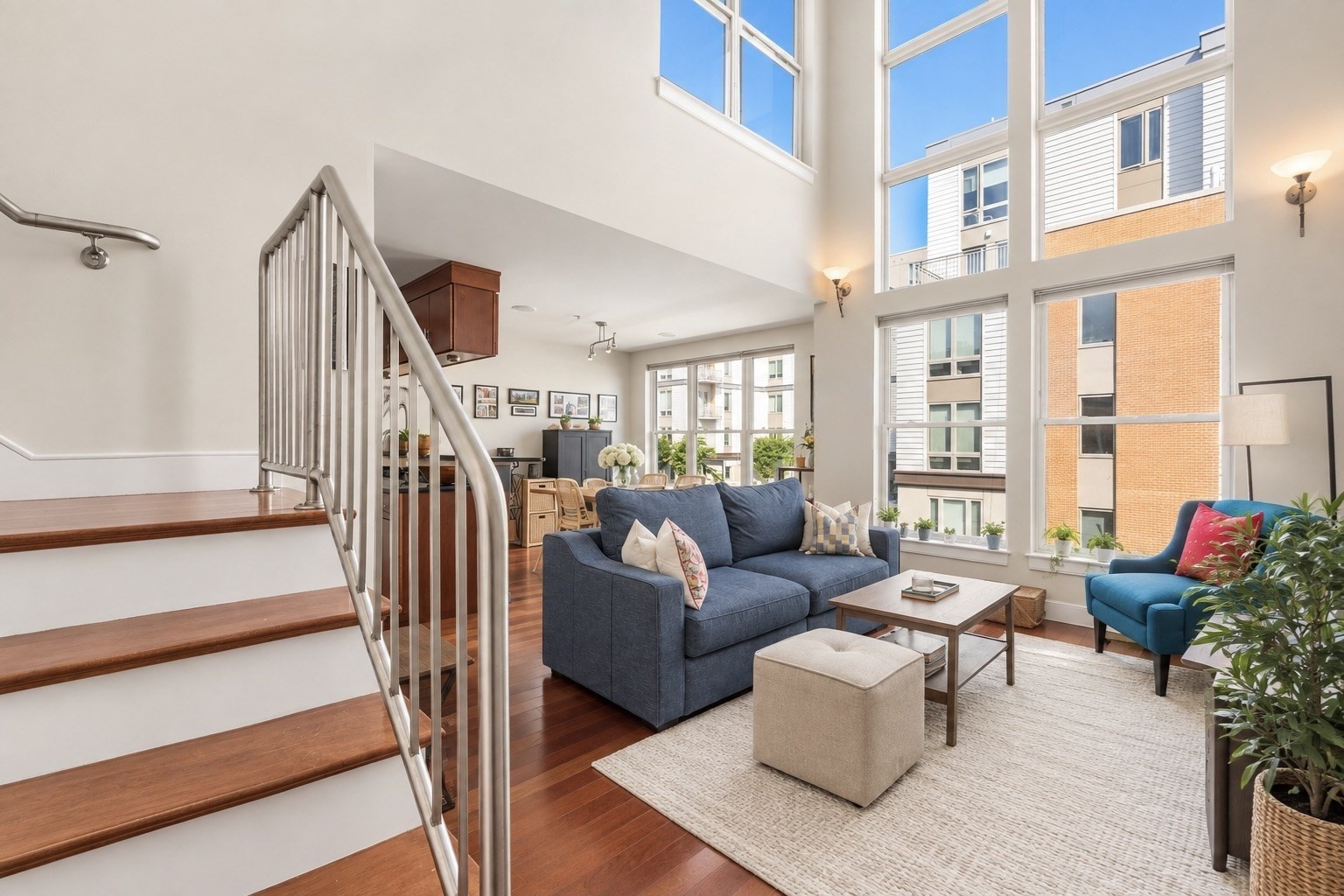 Bright double-height living room with tall windows, a blue sofa, and an open staircase.