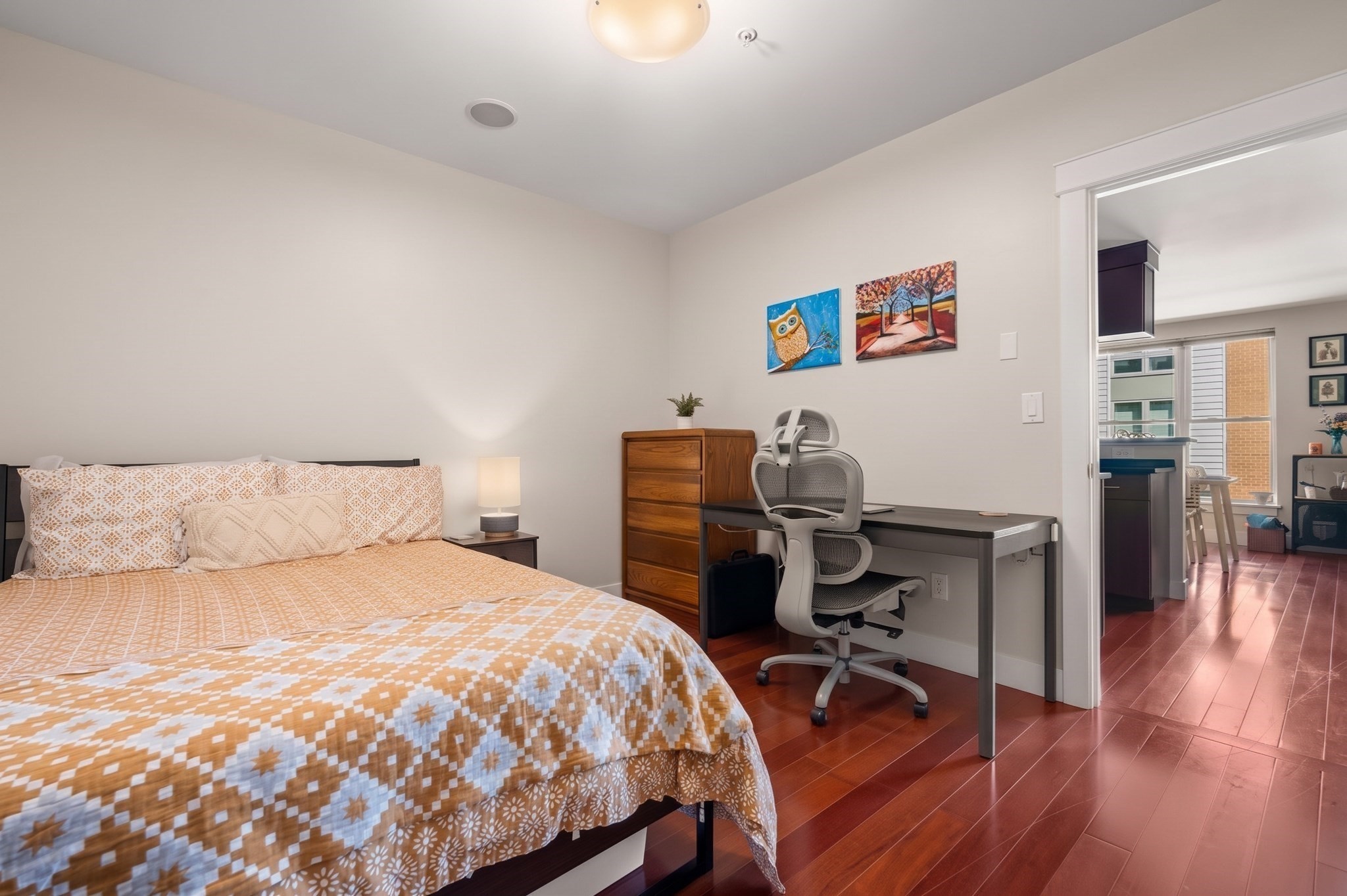 Bedroom with a patterned bedspread, desk and ergonomic chair beside an open doorway to the living area.