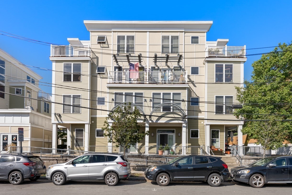 Beige multi-story residential building with balconies and a covered entrance, viewed from the street.