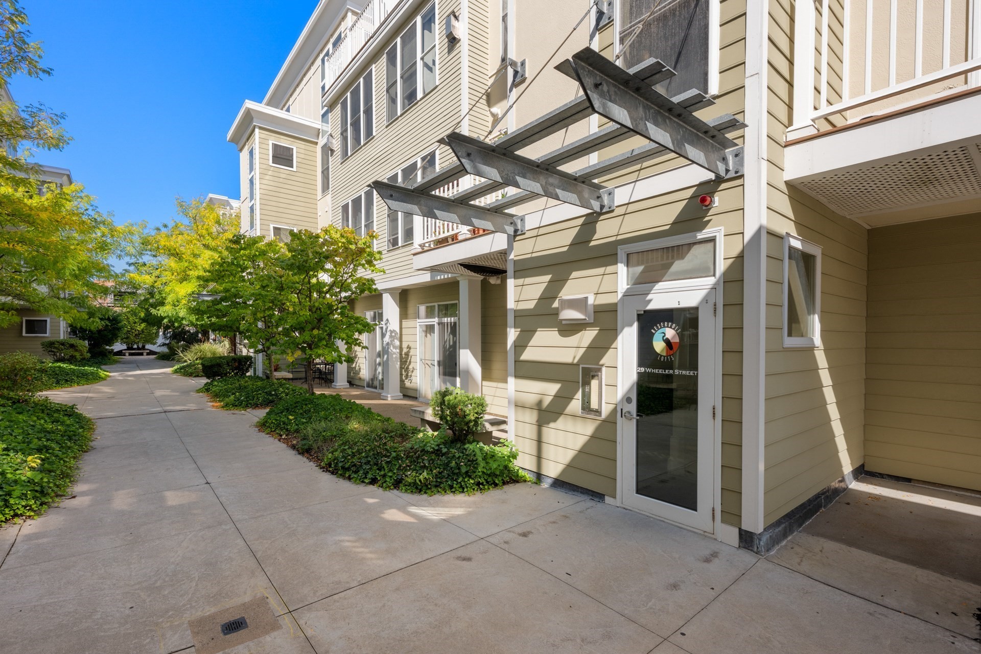 Residential building entrance with a glass door, landscaped walkway, and metal canopy supports.