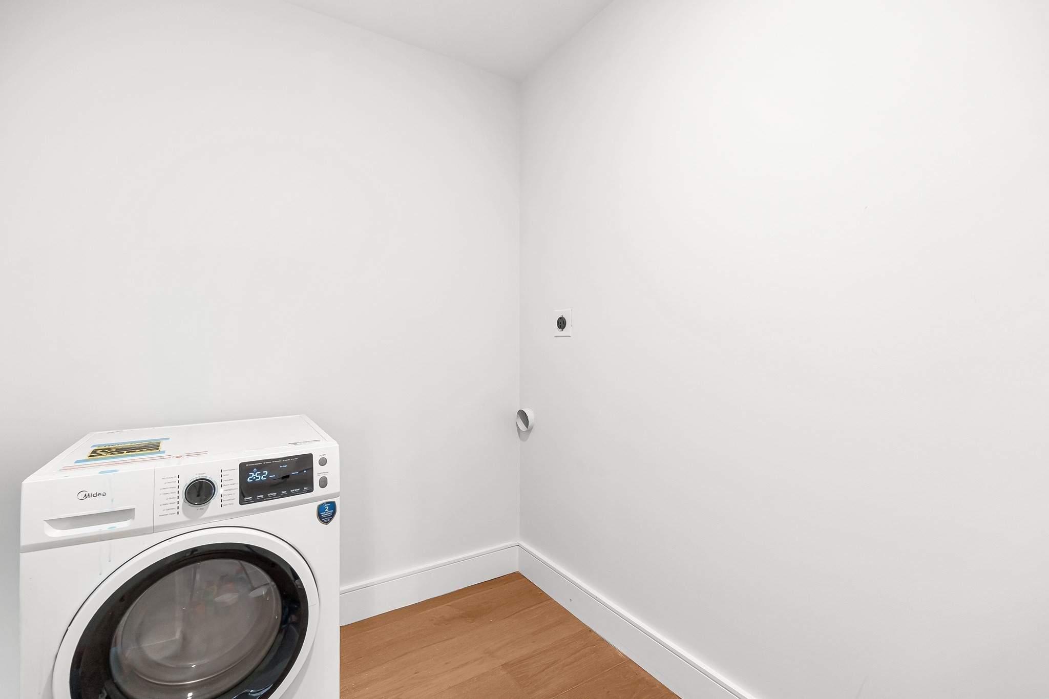 Front-loading washer in a small laundry room with white walls and wood flooring.