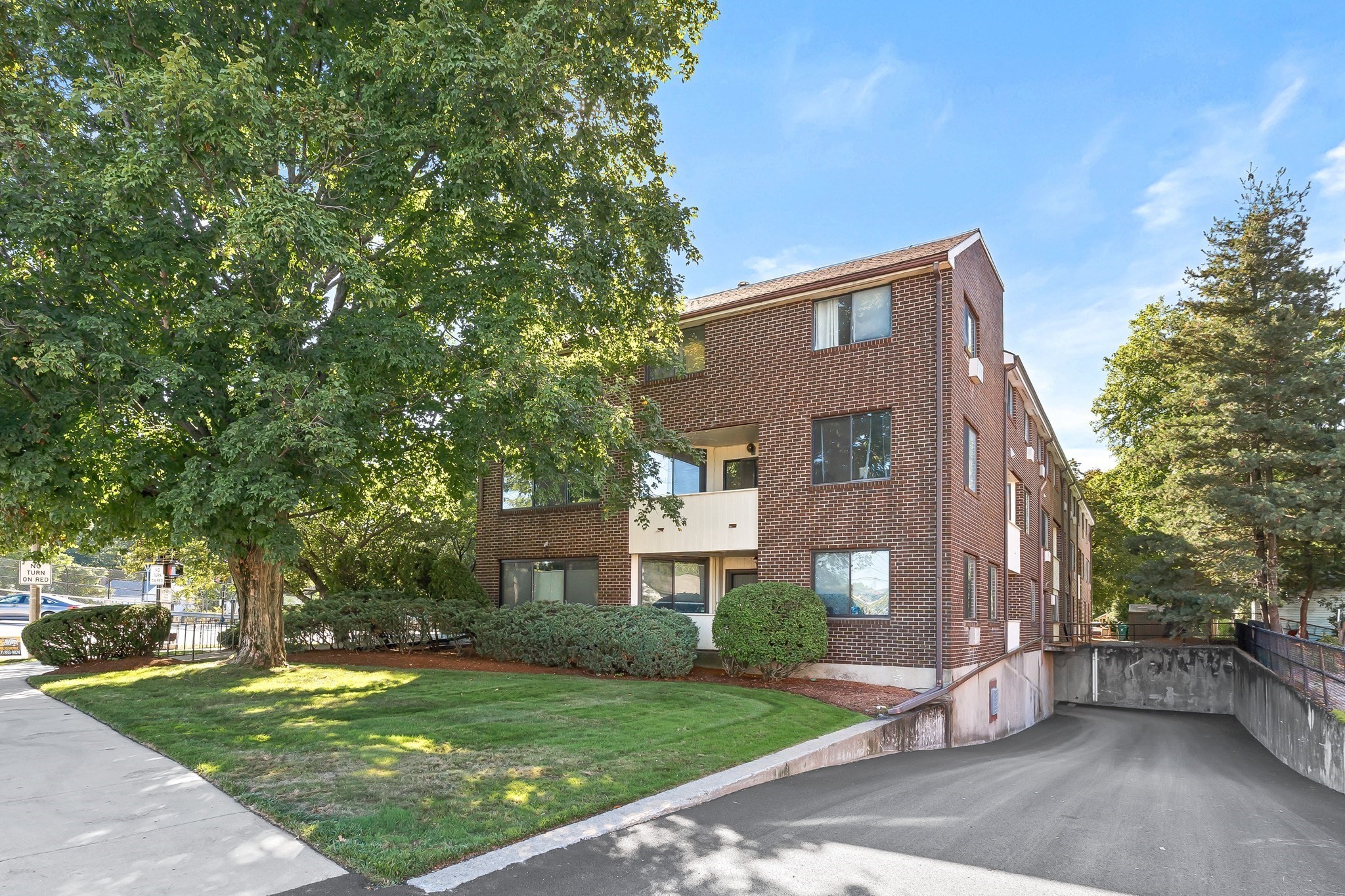 Red-brick apartment building with white balconies, mature trees, and a driveway to the garage.