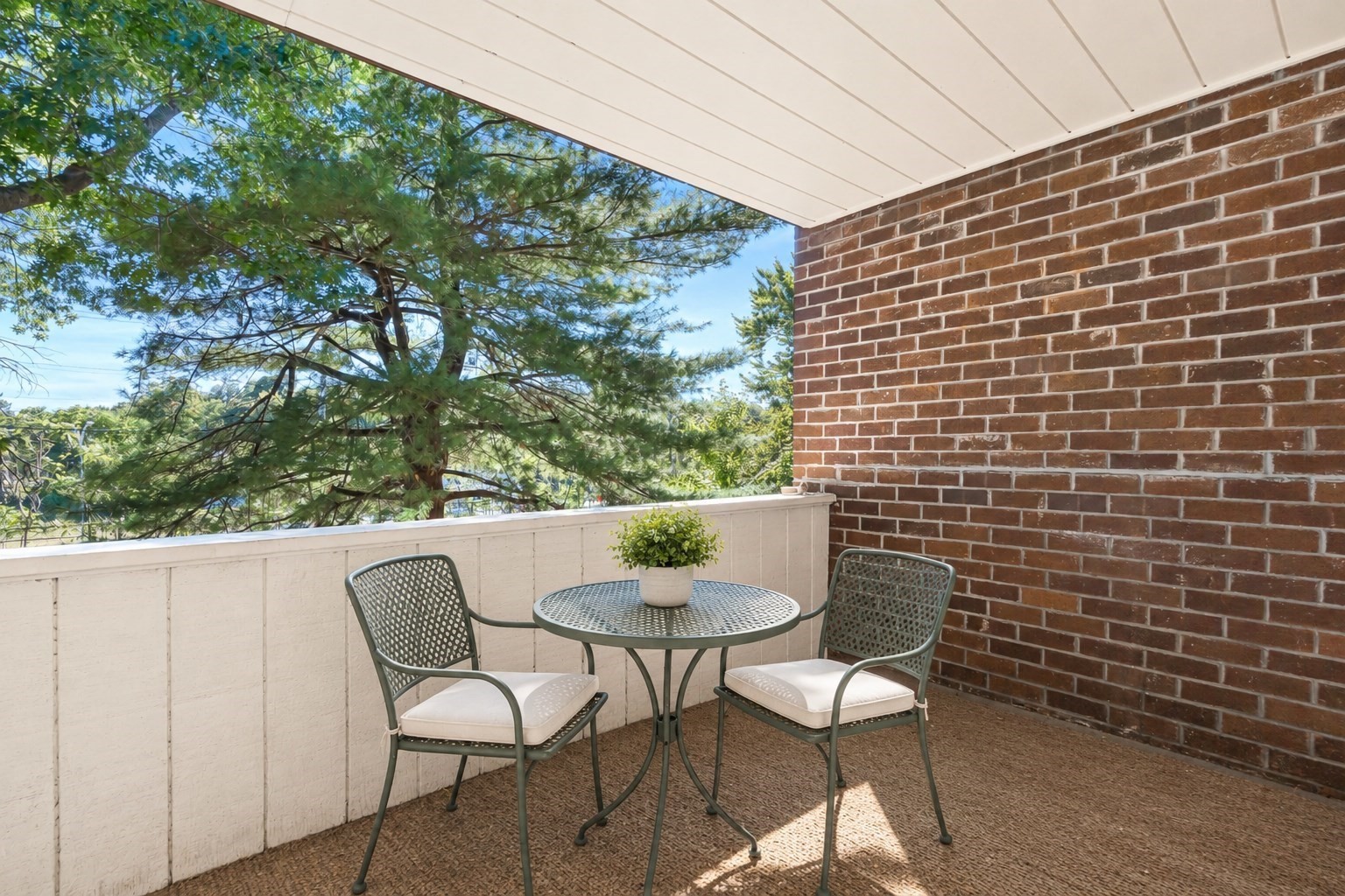 Apartment balcony with a round table, two chairs, and leafy trees beyond the railing.