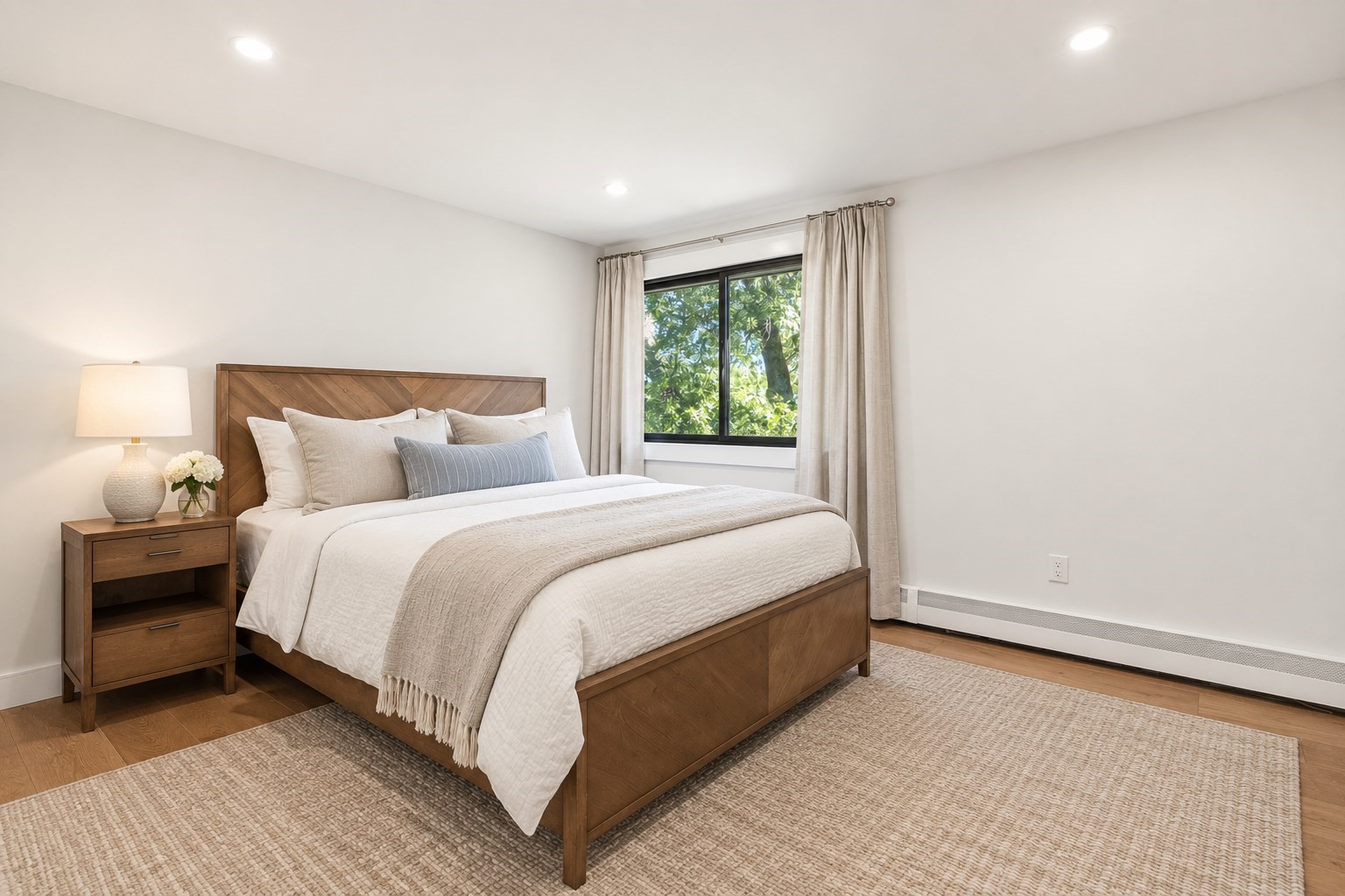 Staged bedroom with a wooden bed, neutral bedding, and a window overlooking leafy trees.