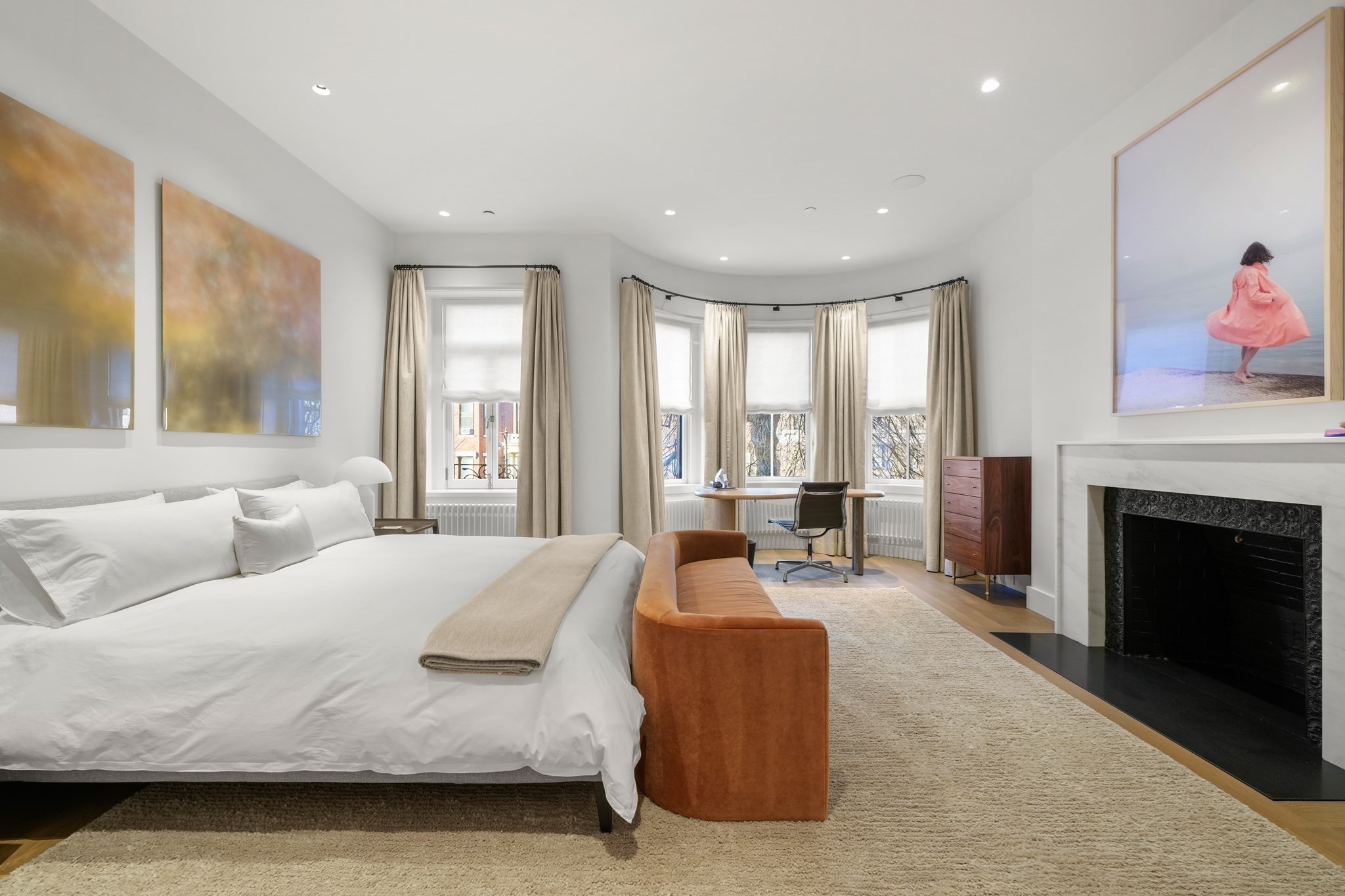 Bright bedroom with a bay window, white bed, rust-colored sofa, and fireplace.