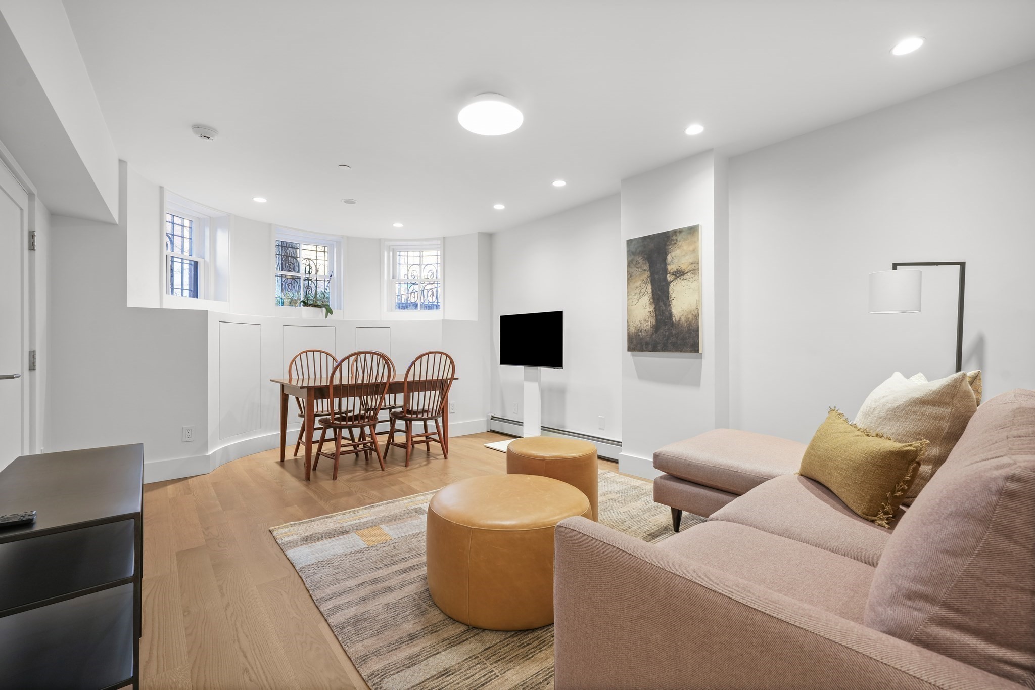 Living and dining area with a blush sectional, round ottomans, and three high windows.