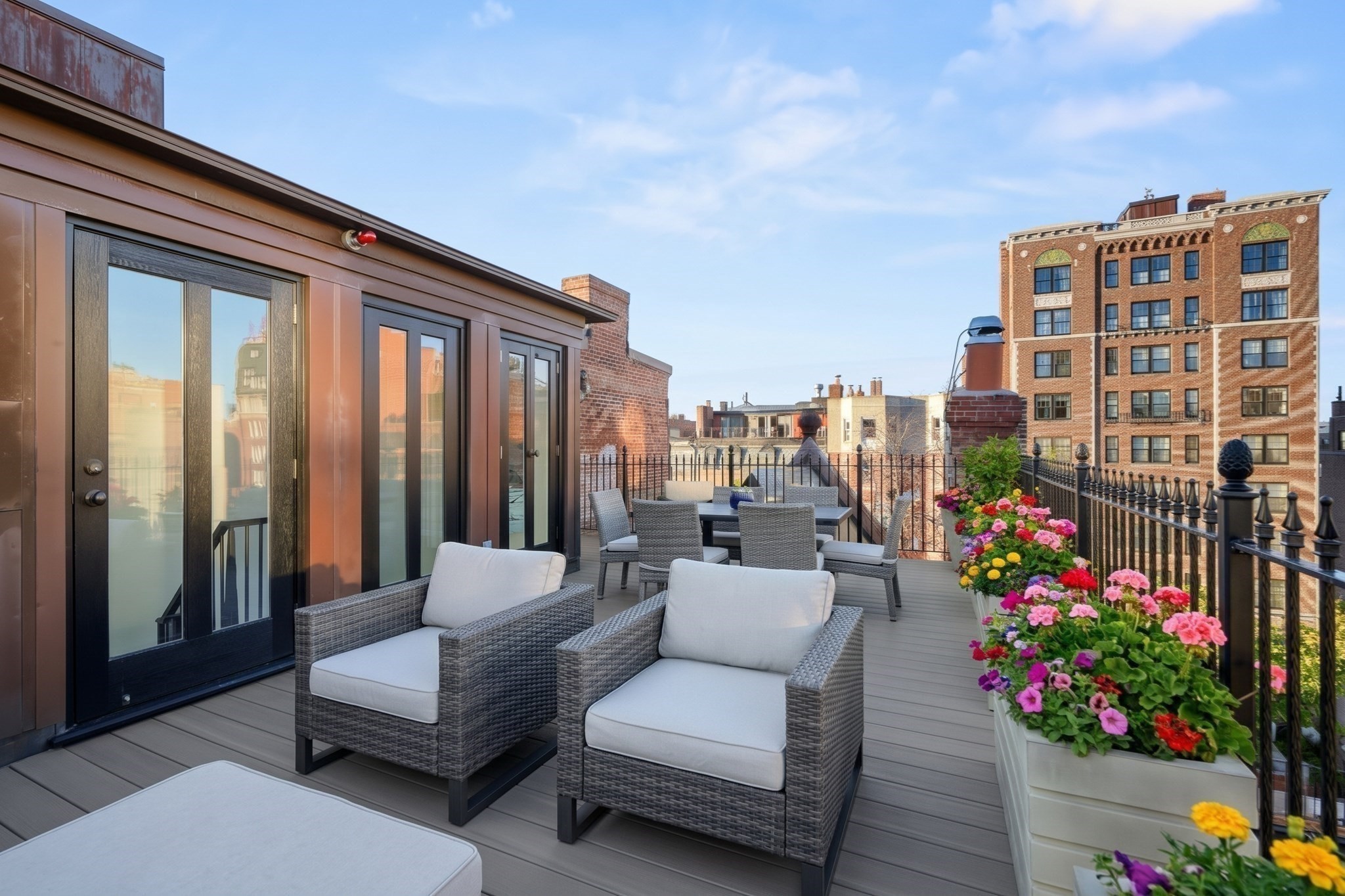 Rooftop terrace with wicker seating and colorful flower planters overlooking brick buildings.