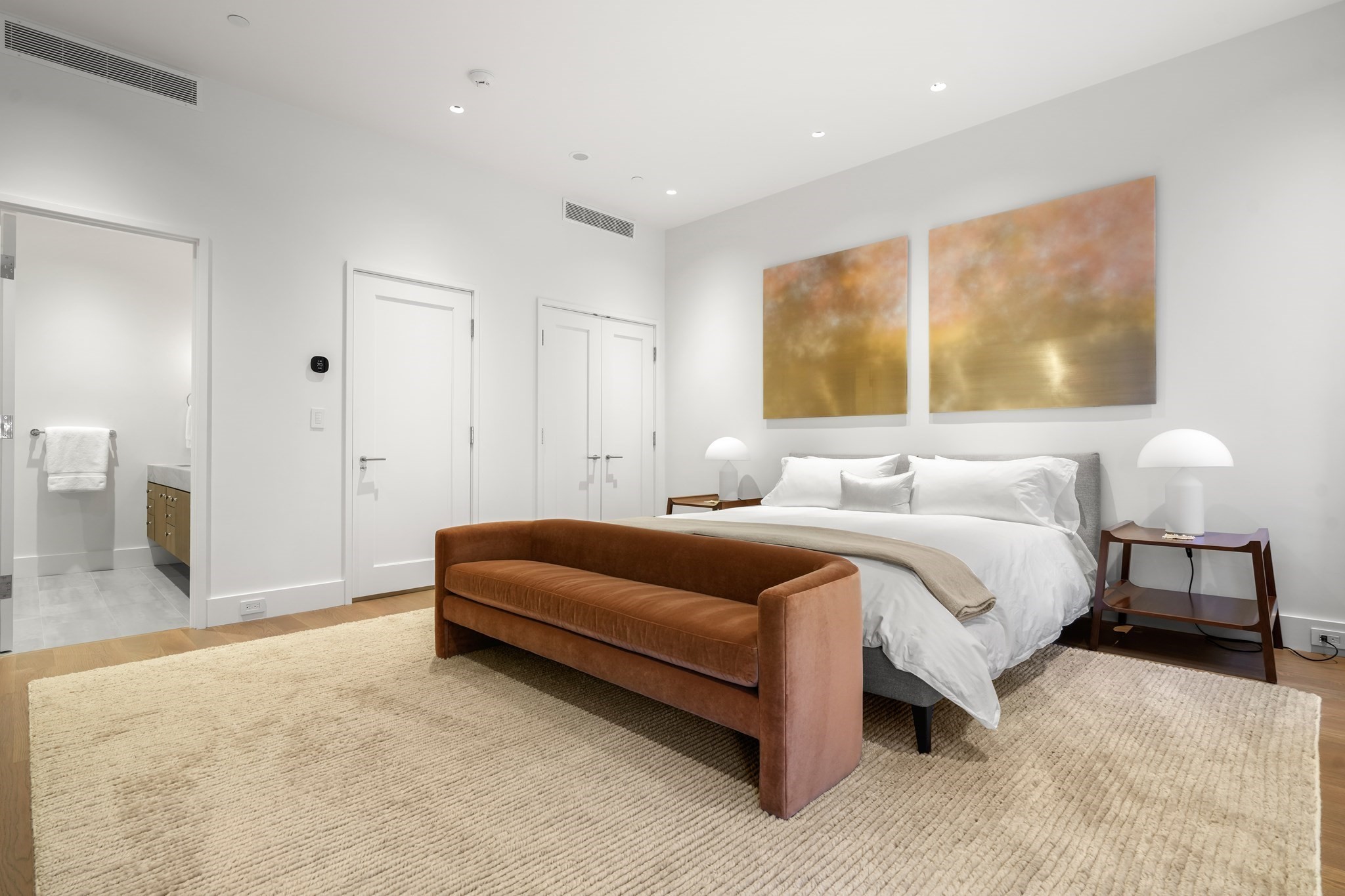 Modern bedroom with a white bed, rust-colored bench, and gold-toned abstract paintings.