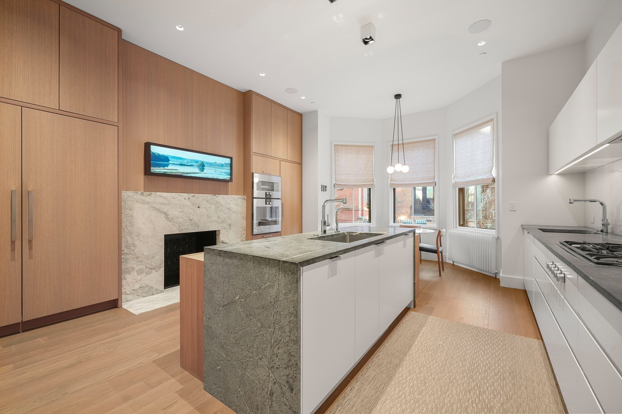 Modern kitchen with a stone island, warm wood cabinetry, and a marble fireplace beside three windows.