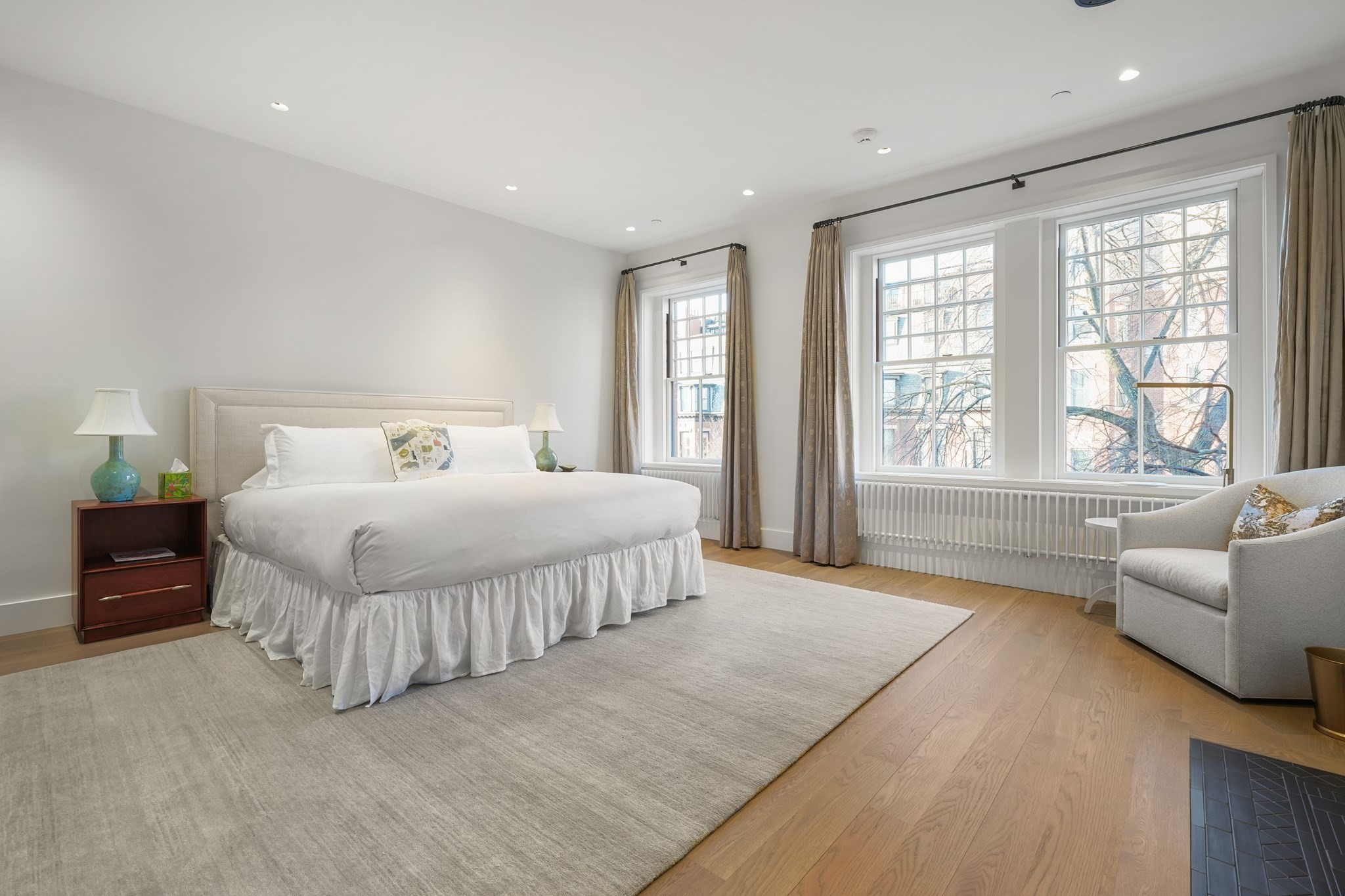 Furnished bedroom with a large white bed, tall multi-pane windows, radiator, and hardwood floors.