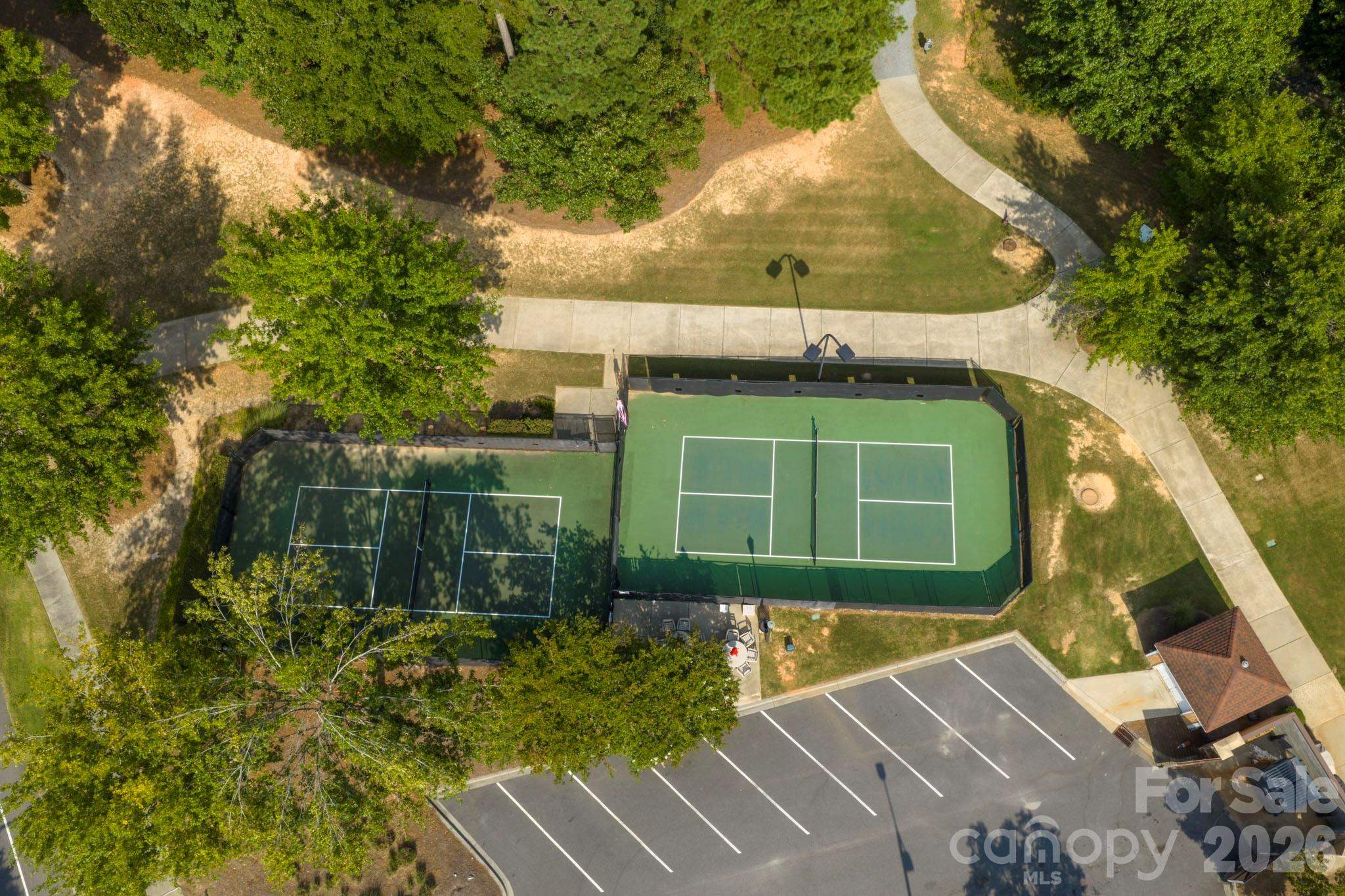 Aerial view of two fenced tennis courts beside a parking lot and tree-lined walkways.