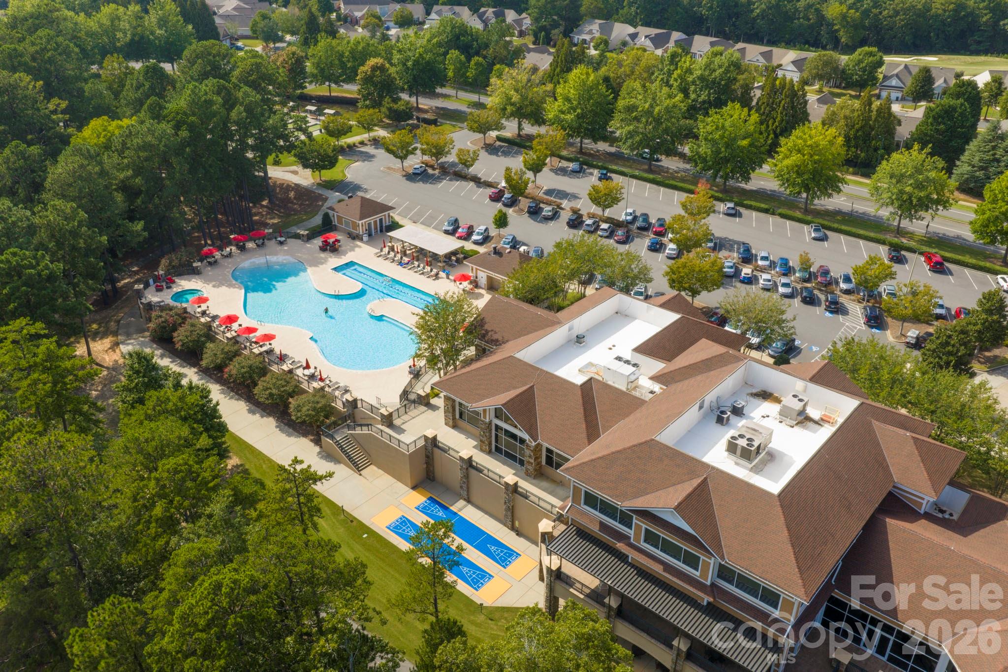 Aerial view of a community pool, clubhouse, and outdoor courts surrounded by trees.