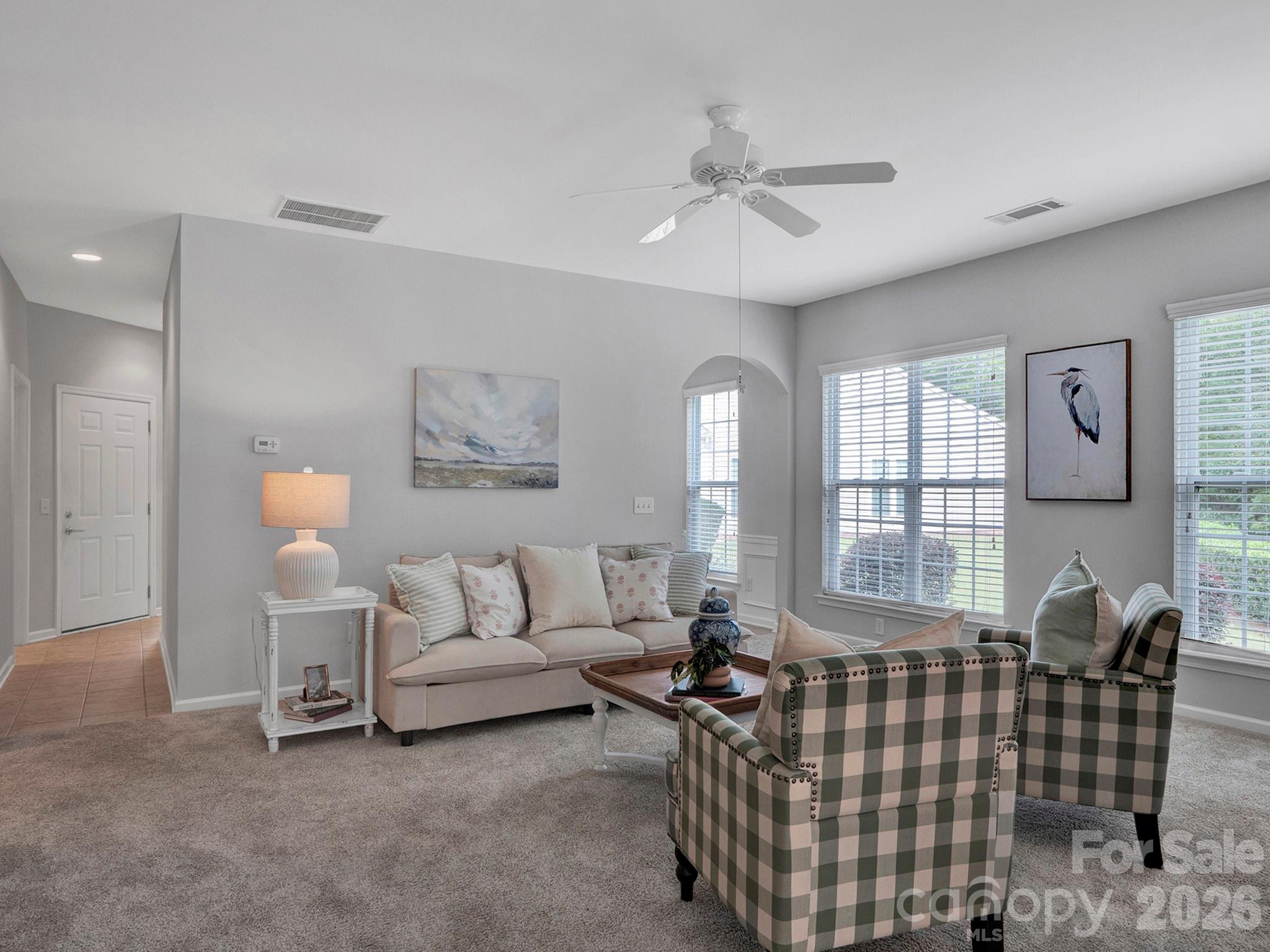 Furnished living room with plaid armchairs, a light sofa, and large windows with blinds.