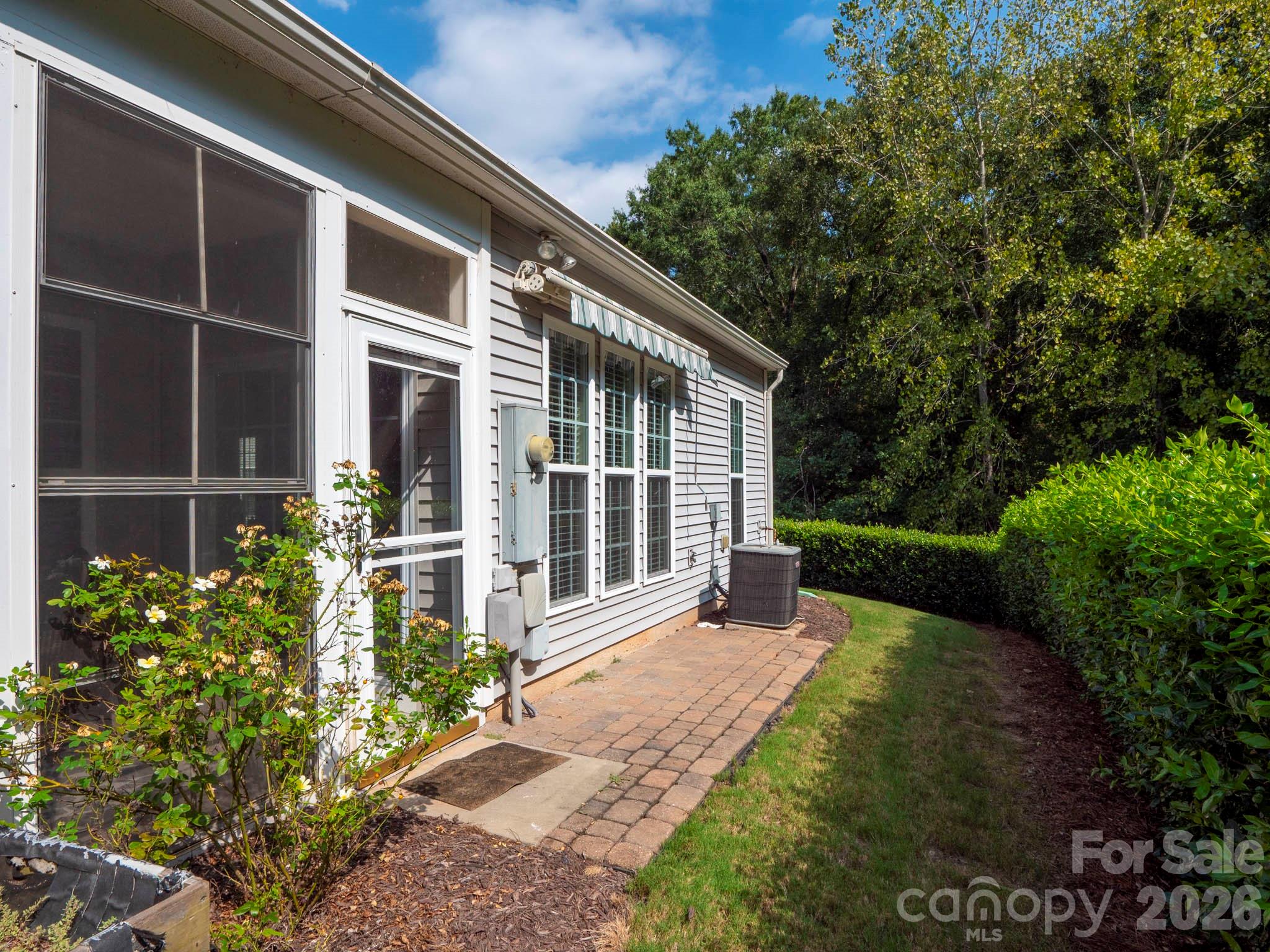 Home’s rear exterior with a screened porch, brick walkway, garden beds, and wooded backdrop.
