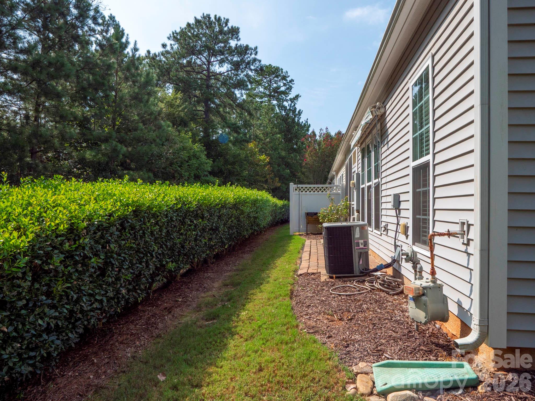 House side yard with a long hedge, grass strip, outdoor HVAC unit, and fenced patio.