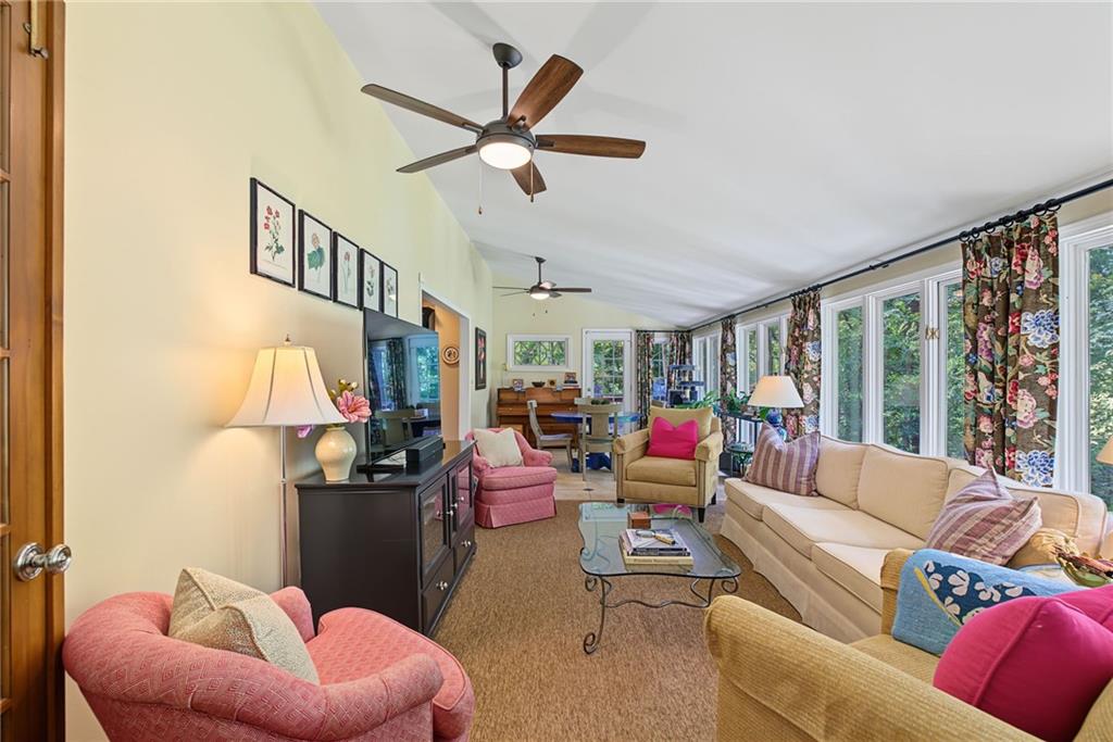 Furnished living room with vaulted ceiling, large windows overlooking trees, and colorful seating.