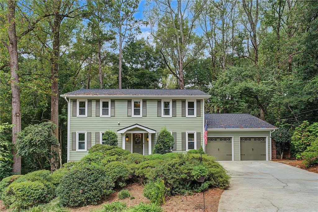 Two-story pale green house with an attached two-car garage, surrounded by mature trees.