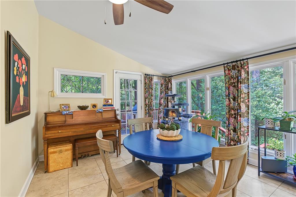Dining room with a blue round table, upright piano, and large windows overlooking greenery.