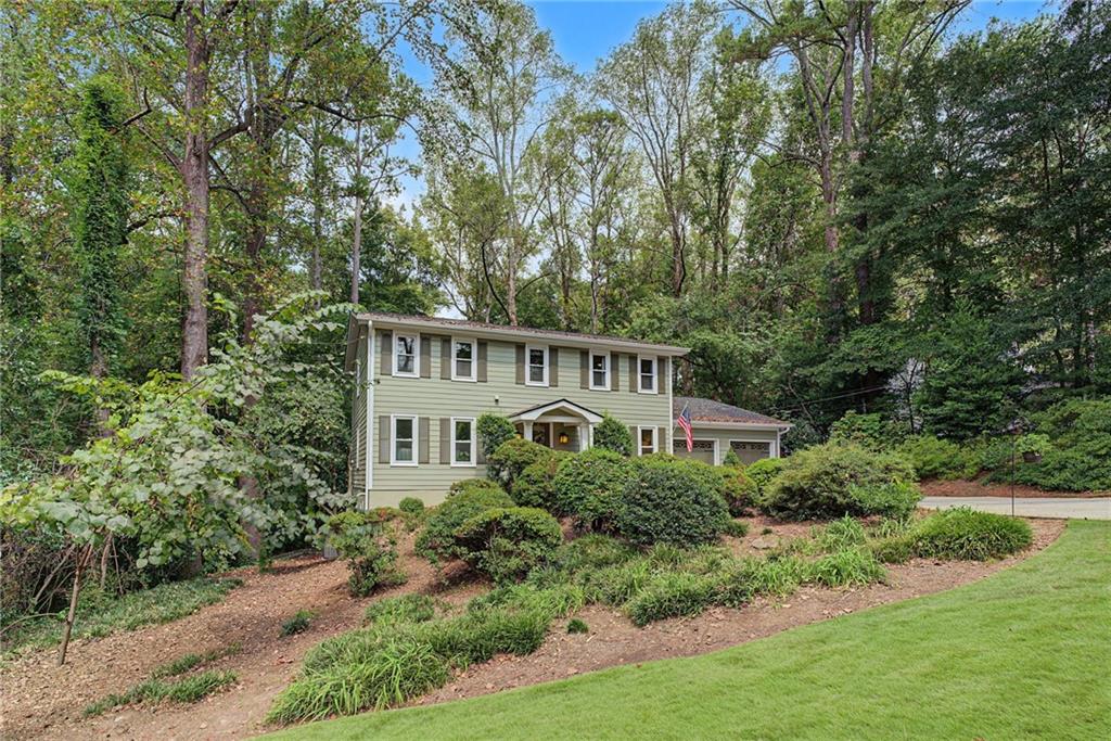 Two-story green house with an attached garage, landscaped shrubs, and mature trees.