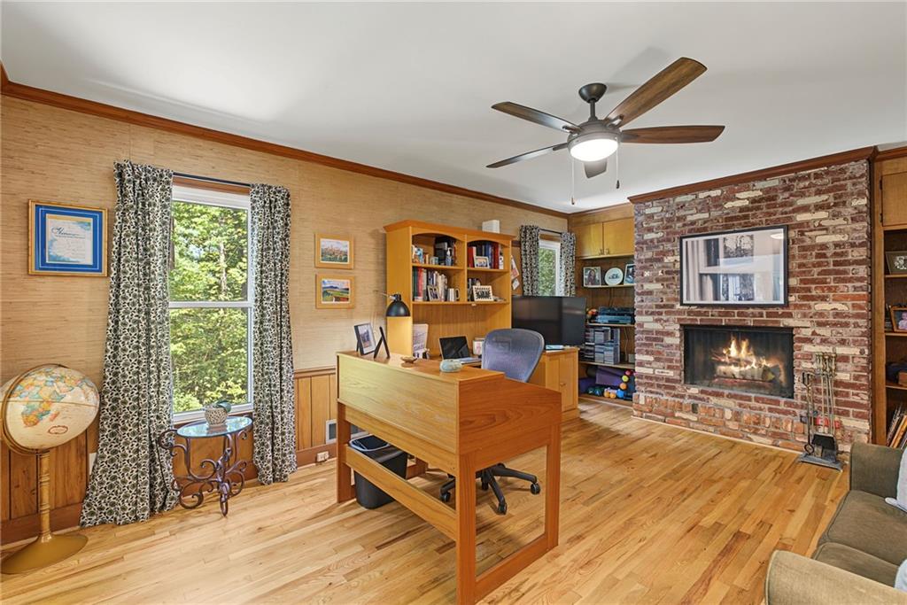 Furnished home office with a wooden desk, brick fireplace, and windows overlooking greenery.