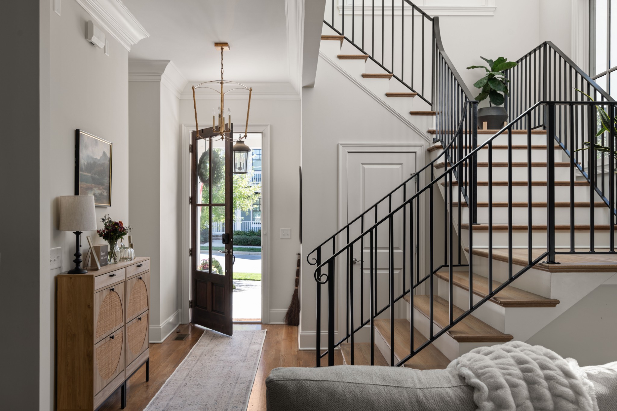 Bright entry foyer with an open front door, wood floors, and a black-railed staircase.
