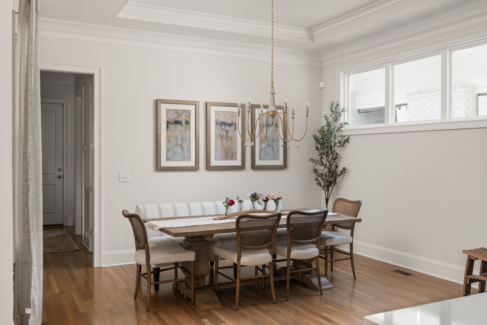 Dining room with a long wooden table, upholstered chairs, and a brass chandelier beneath bright windows.