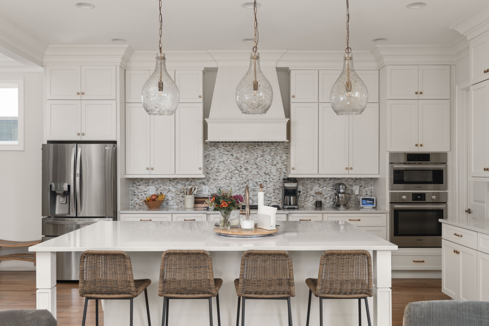 Bright white kitchen with a large island, woven bar stools, pendant lights, and stainless steel appliances.