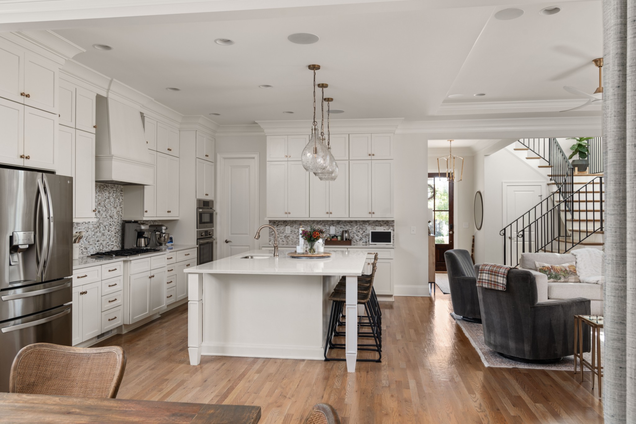 Bright white kitchen with a large island opens to a furnished living area and staircase.