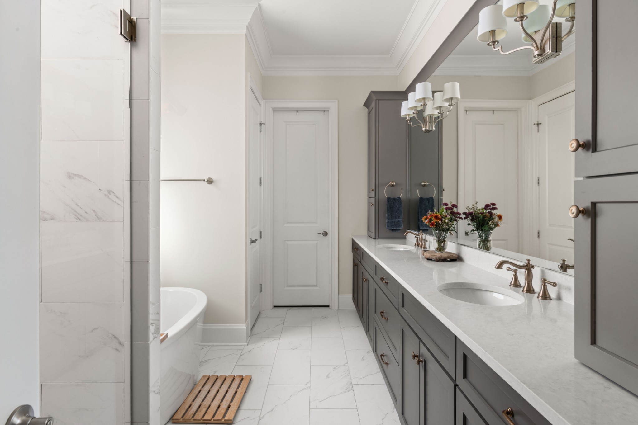 Bathroom with a double vanity, gray cabinetry, marble-look tile, and a freestanding tub.