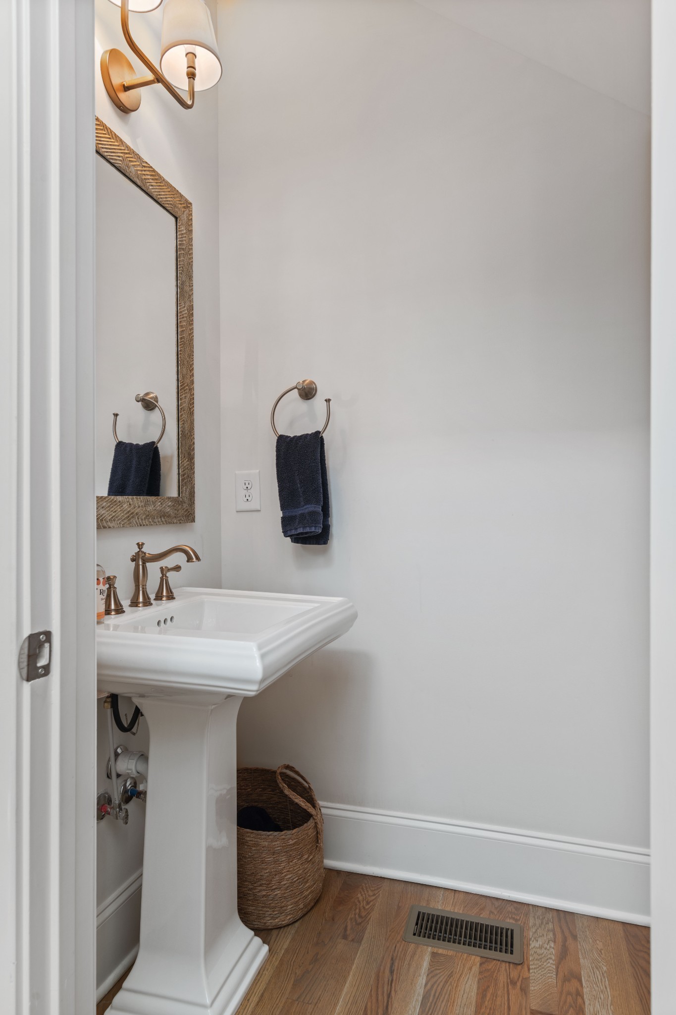 Powder room with a white pedestal sink, framed mirror, brass sconces, and hardwood flooring.
