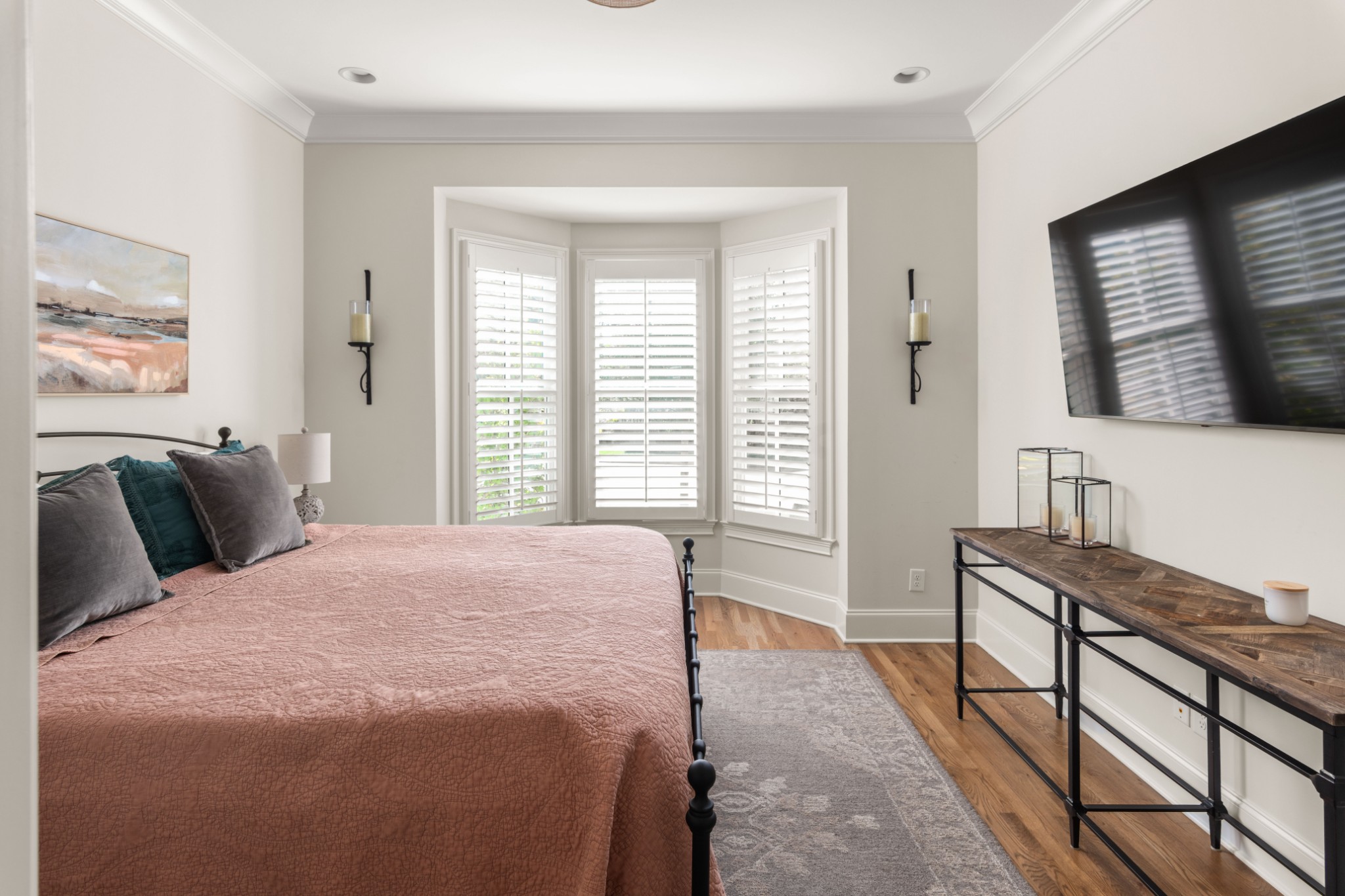 Staged bedroom with a bay window, plantation shutters, and a large wall-mounted television.