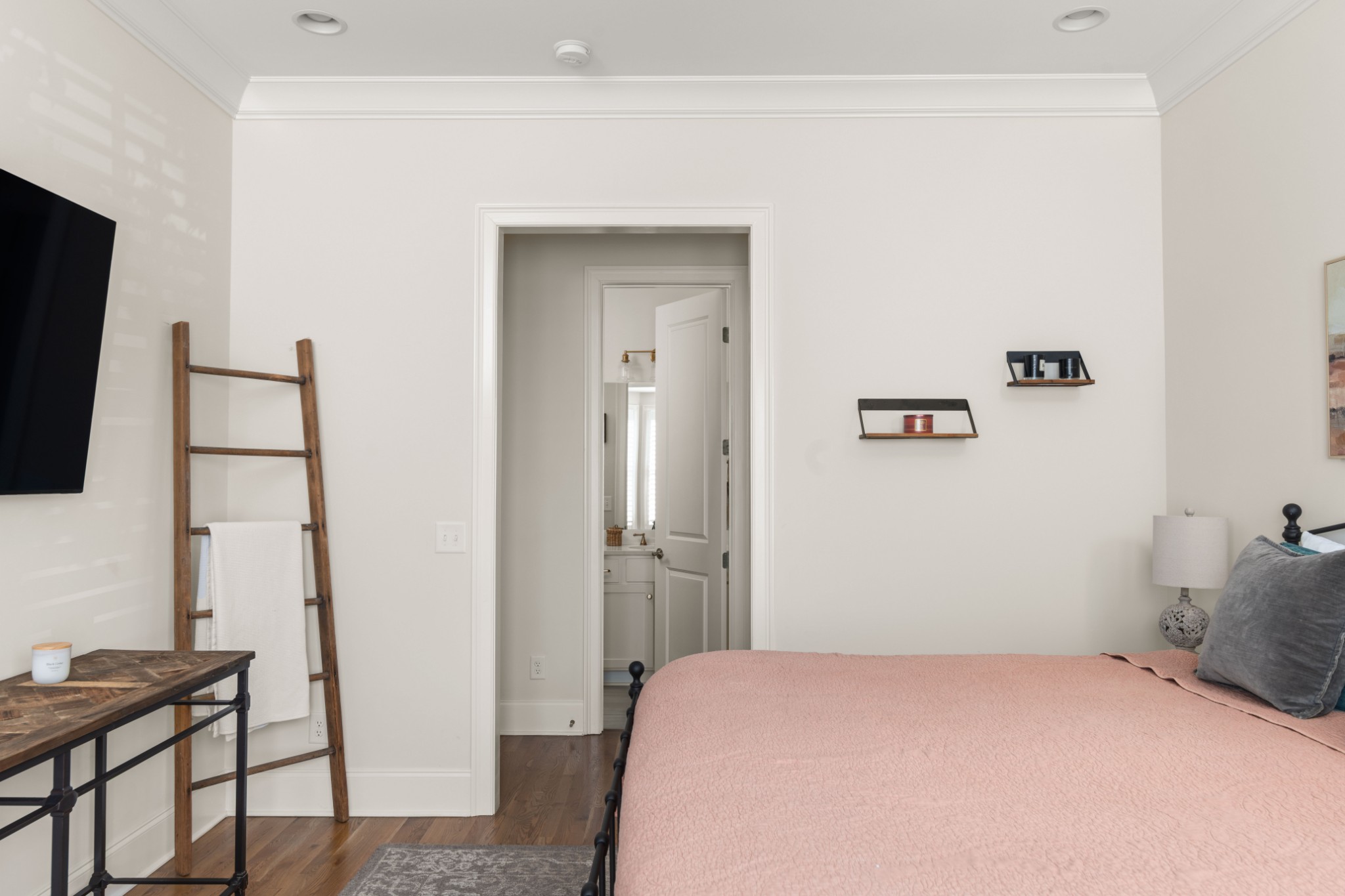 Bedroom with a pink quilt, wooden towel ladder, and open doorway to an ensuite bathroom.