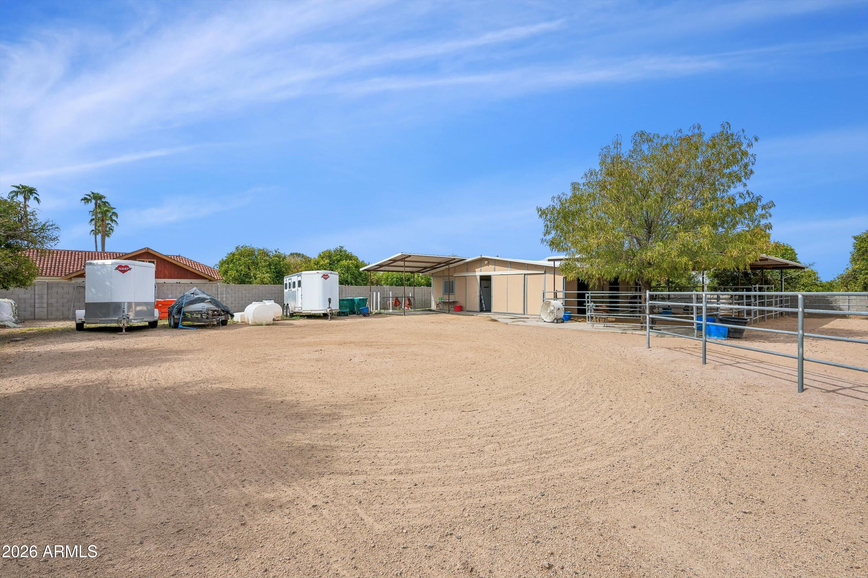 Sandy property yard with fenced animal pens, a shade tree, and trailers beside a metal-roofed outbuilding.