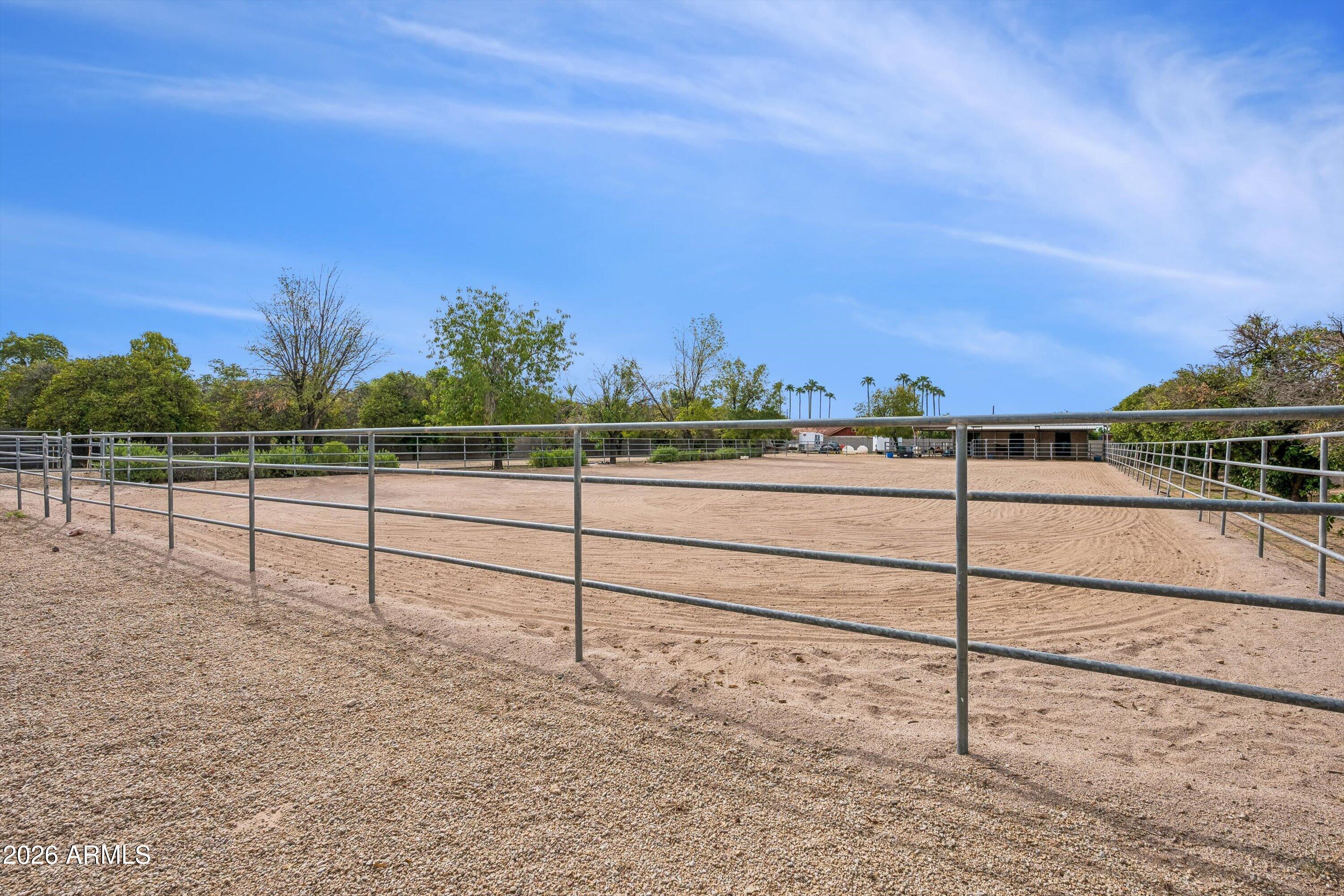 Fenced sandy riding arena with a stable building and trees in the background.