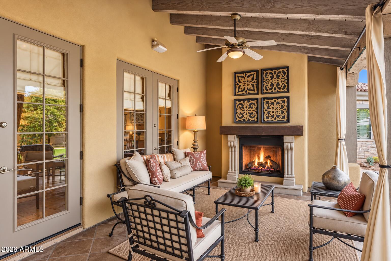 Covered patio with a lit fireplace, exposed wood beams, and cushioned seating.