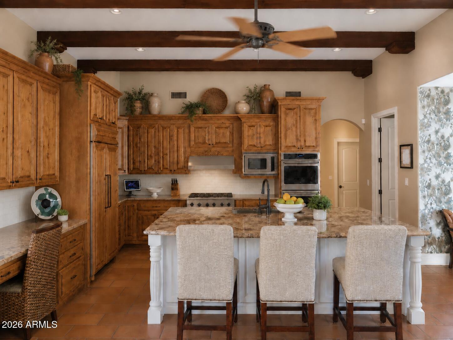 Kitchen with honey-toned cabinetry, exposed beams, and a large marbled island with three bar stools.