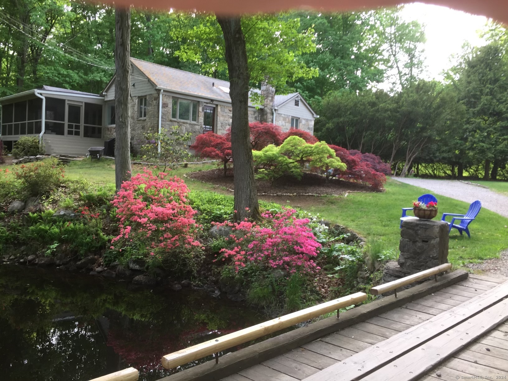 Stone-sided home behind flowering gardens, with a screened porch, pond, and wooden footbridge.