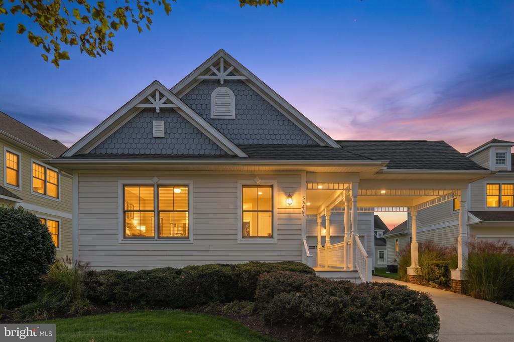 Gray shingle-sided home with a covered porch, glowing windows, and landscaped front yard at dusk.