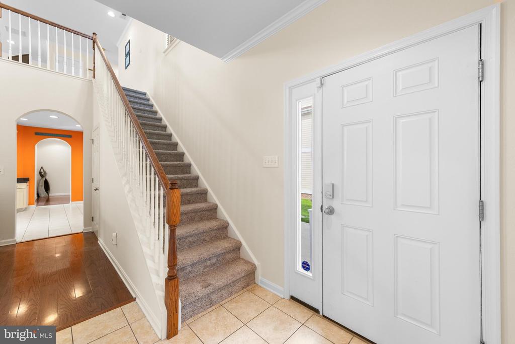 Entry foyer with a white front door, tiled floor, and carpeted staircase beside a hardwood hallway.