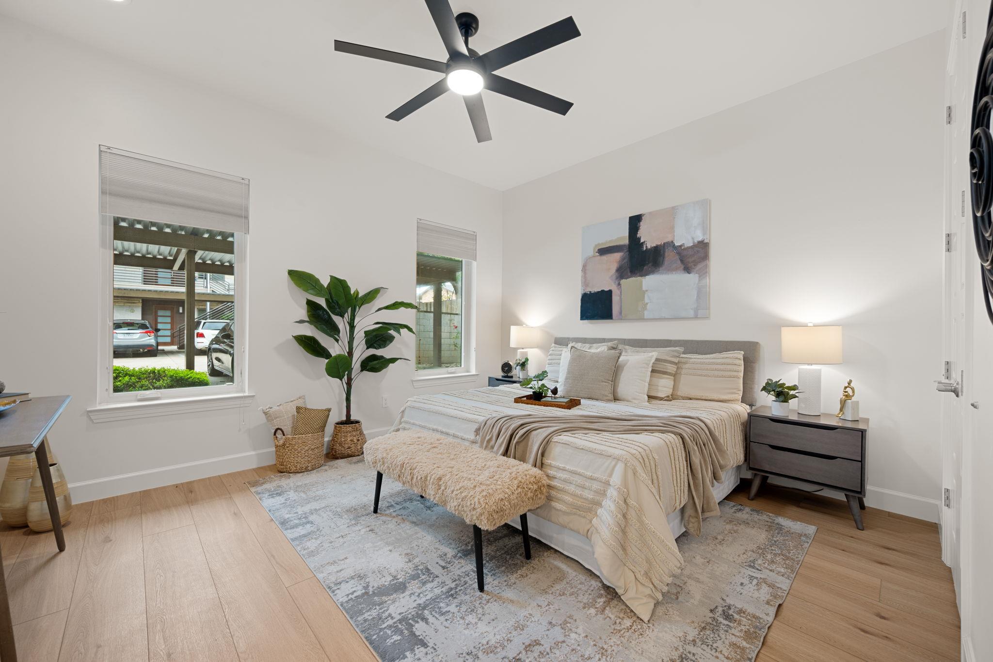 Staged bedroom with a layered neutral bed, abstract art, and black ceiling fan.