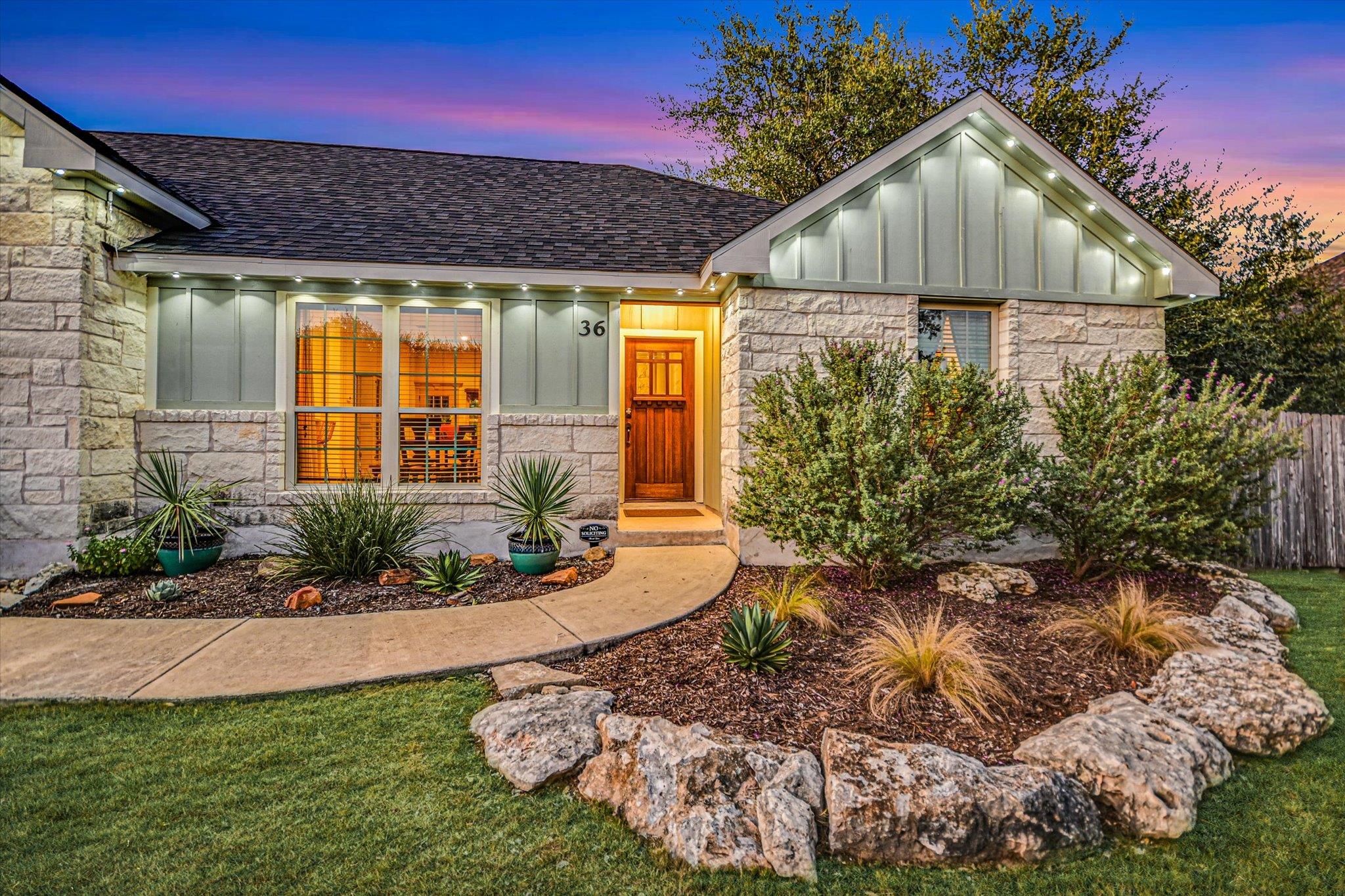 Stone-front home at dusk with a brightly lit wooden entry and landscaped garden beds.