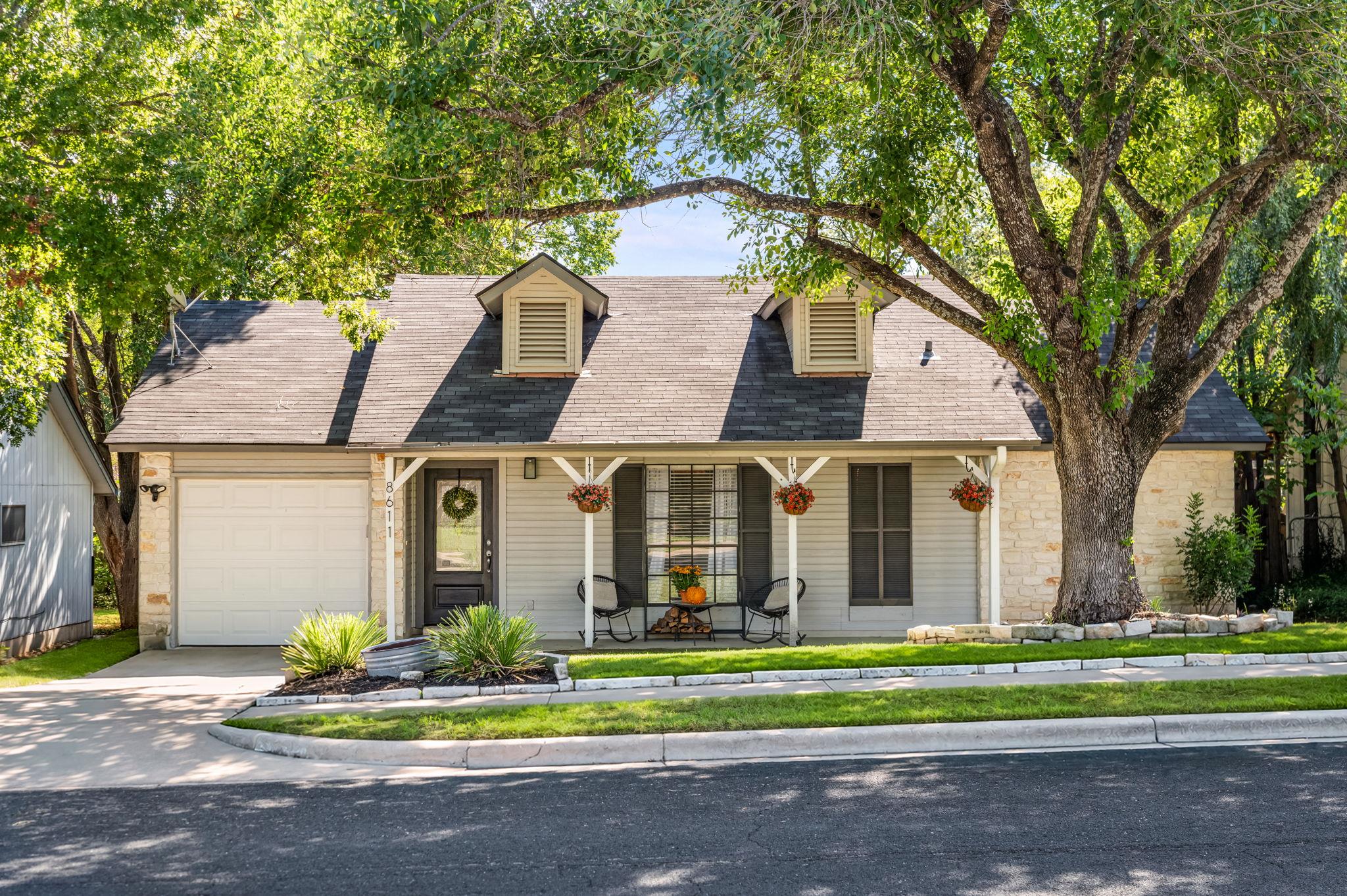 House with stone accents, two dormers, covered porch, attached garage, and a large shade tree.